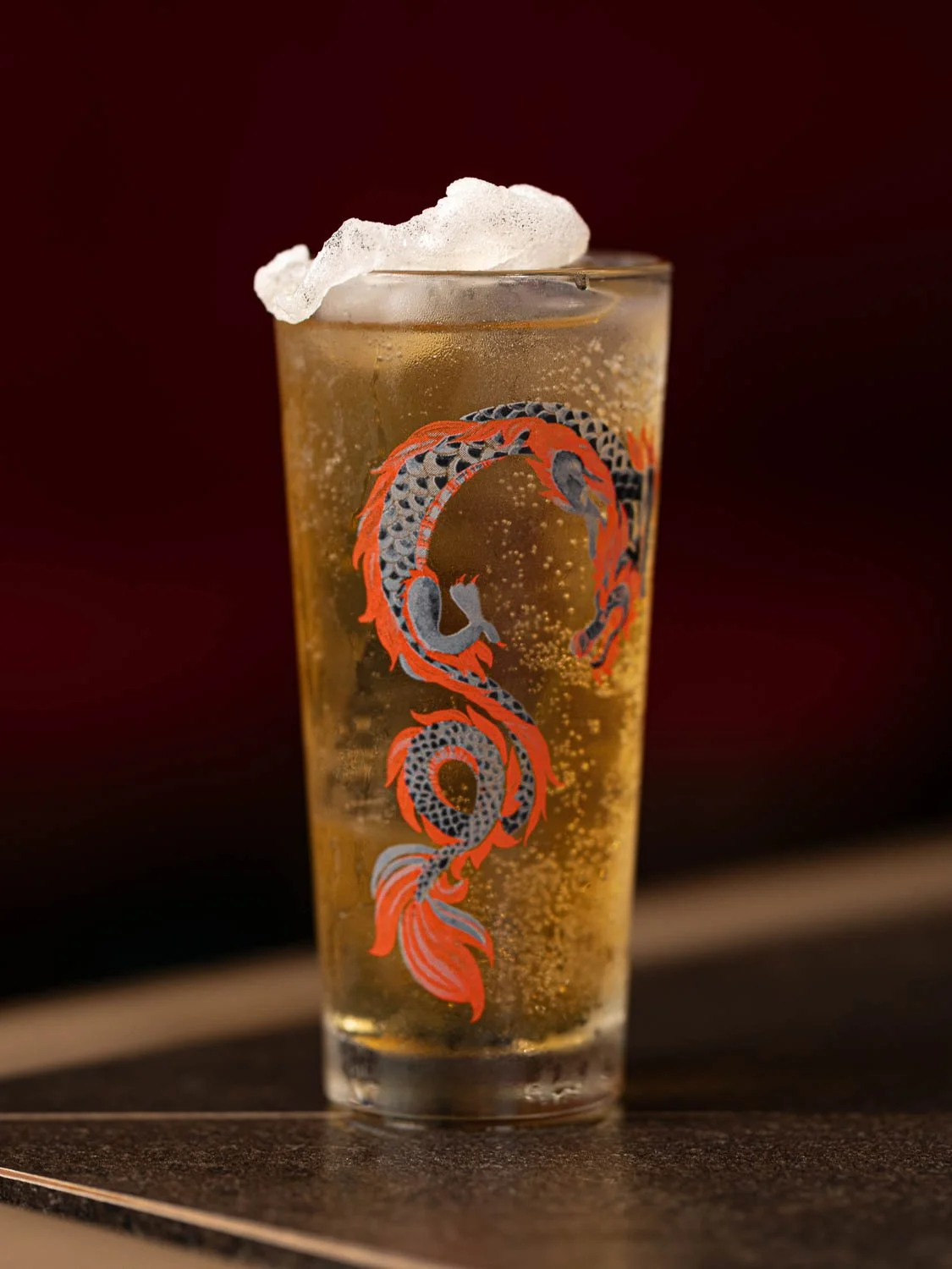 Ein goldfarbener Cocktail in einem hohen Glas, das mit einem japanischen Drachen bedruckt ist, garniert mit einem weißen Chip