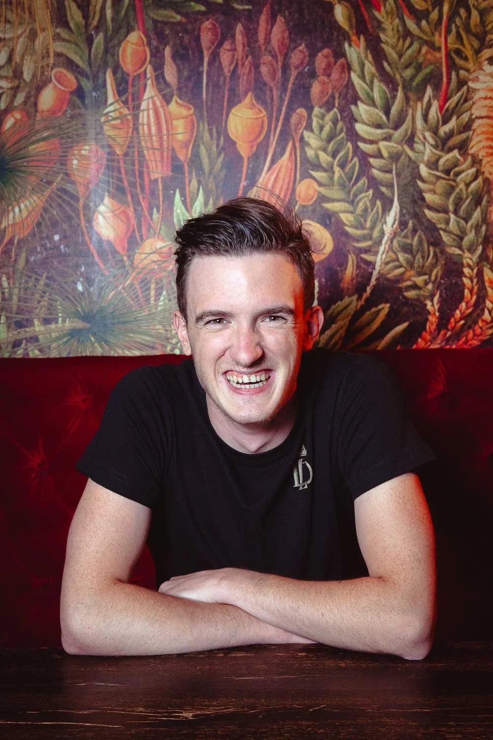 Barkeeper Martin Šiprak sitzt auf einer roten mit Samt bezogenen Bank in der Cocktailbar Liquid Diary und lächelt