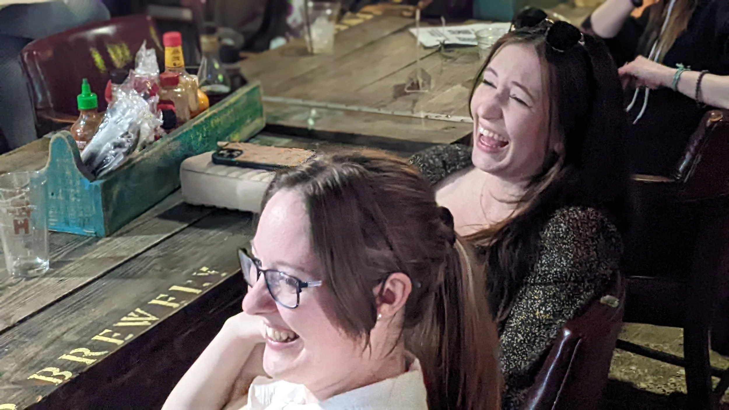 Two women laughing and smiling at a wooden table in a bar or restaurant, with one wearing glasses and the other with sunglasses on her head.