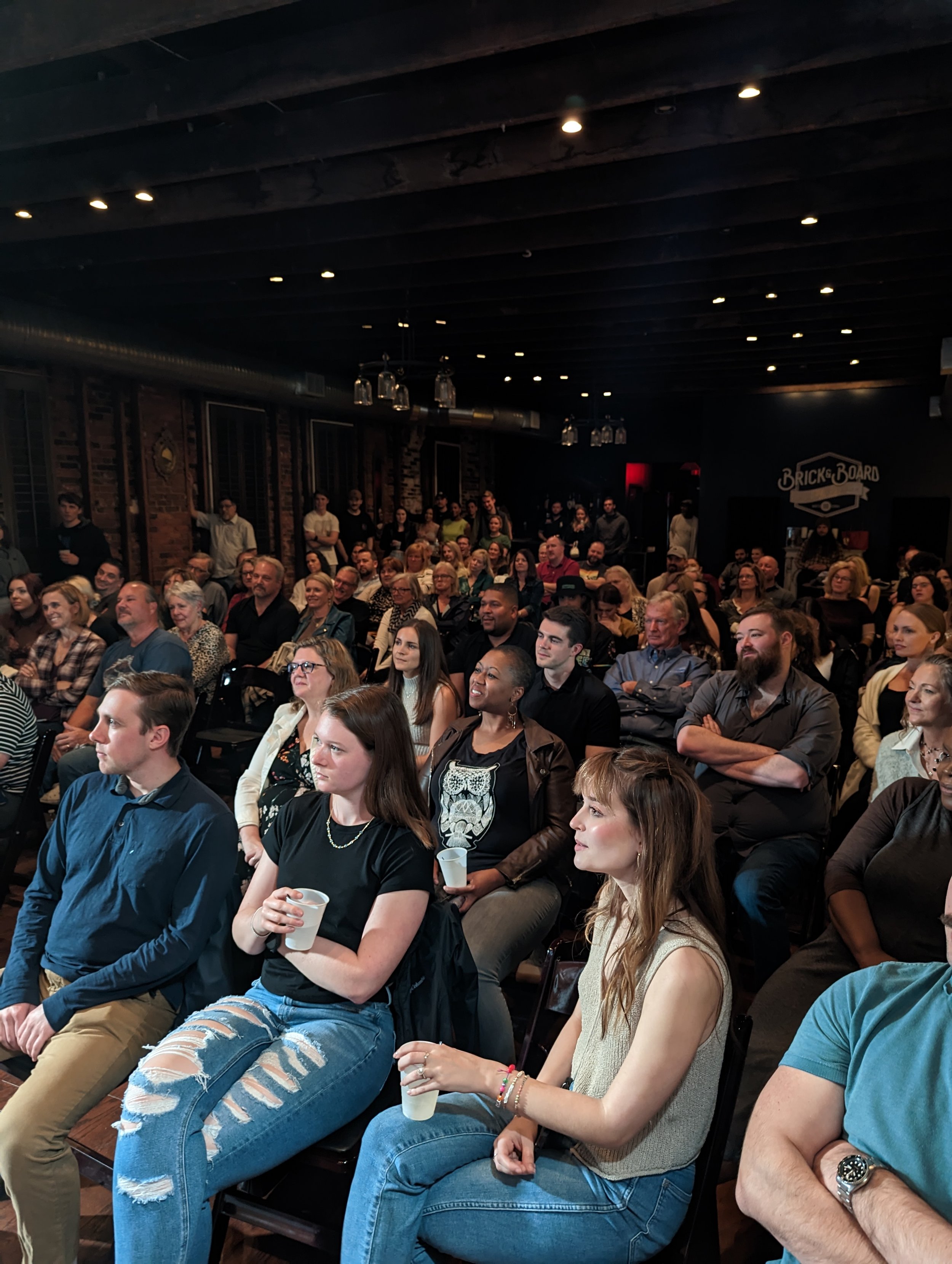 An audience seated in a dimly lit venue, attentively watching a performance or presentation. The room has brick walls with windows and hanging glasses, and a sign that reads 'Brick + Board' is visible in the background.