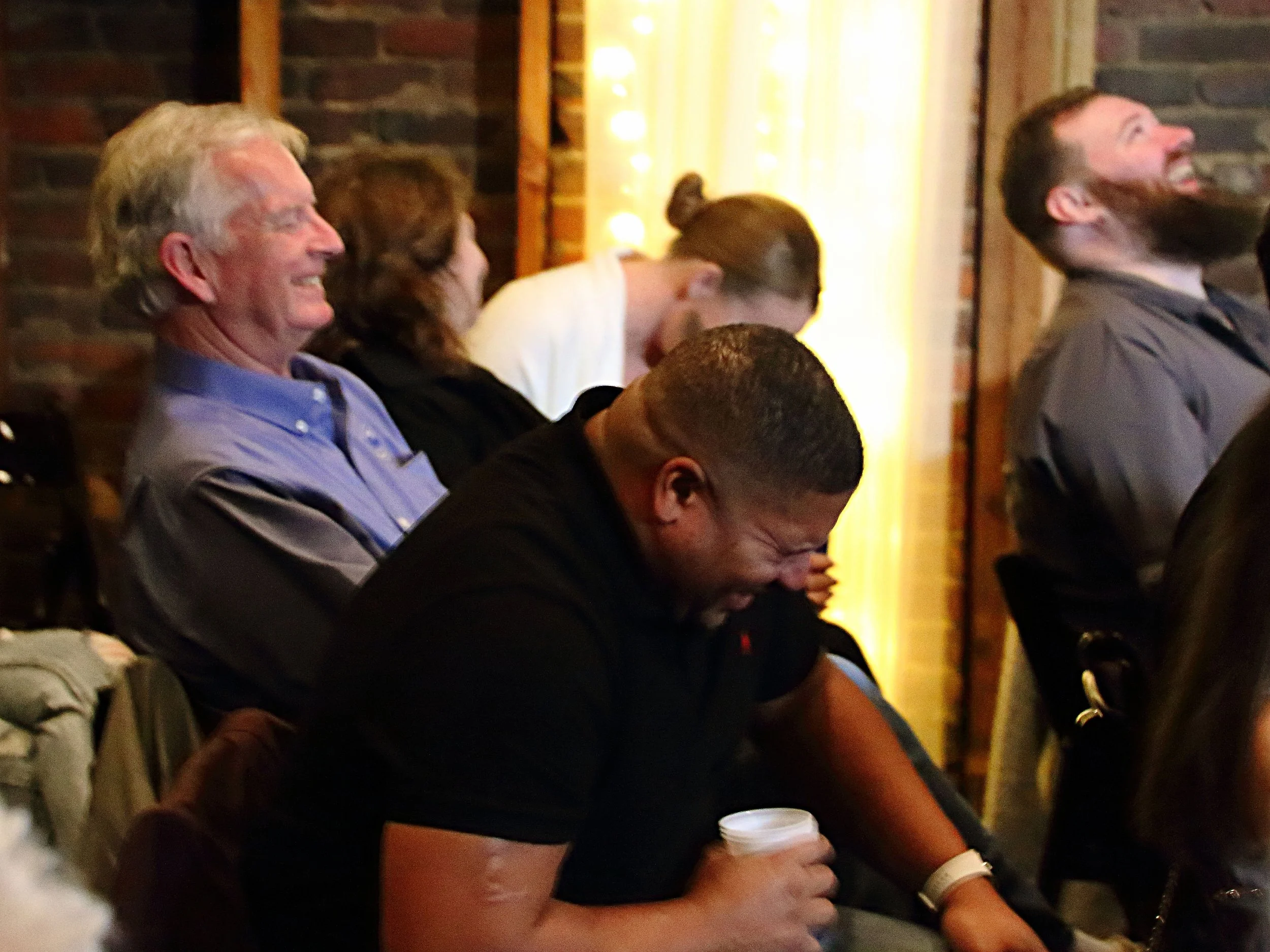 People sitting at a row of chairs, smiling and enjoying a lively event or performance. The background includes a brick wall and decor with warm lighting.