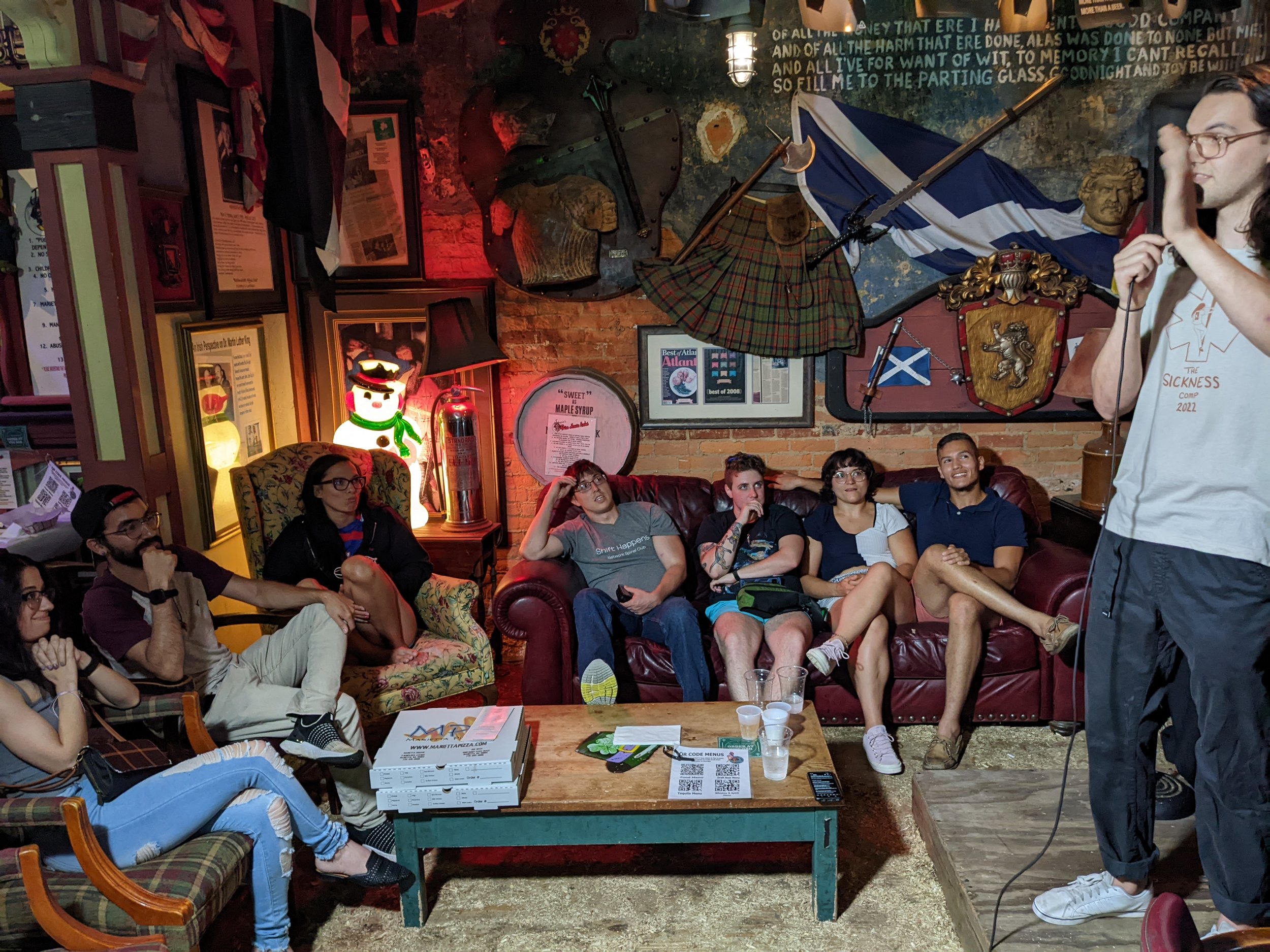 A group of people sitting on a sofa and chairs in a cozy, decorated room with brick walls, artwork, and Scottish-themed decorations, listening to a person standing with a microphone.