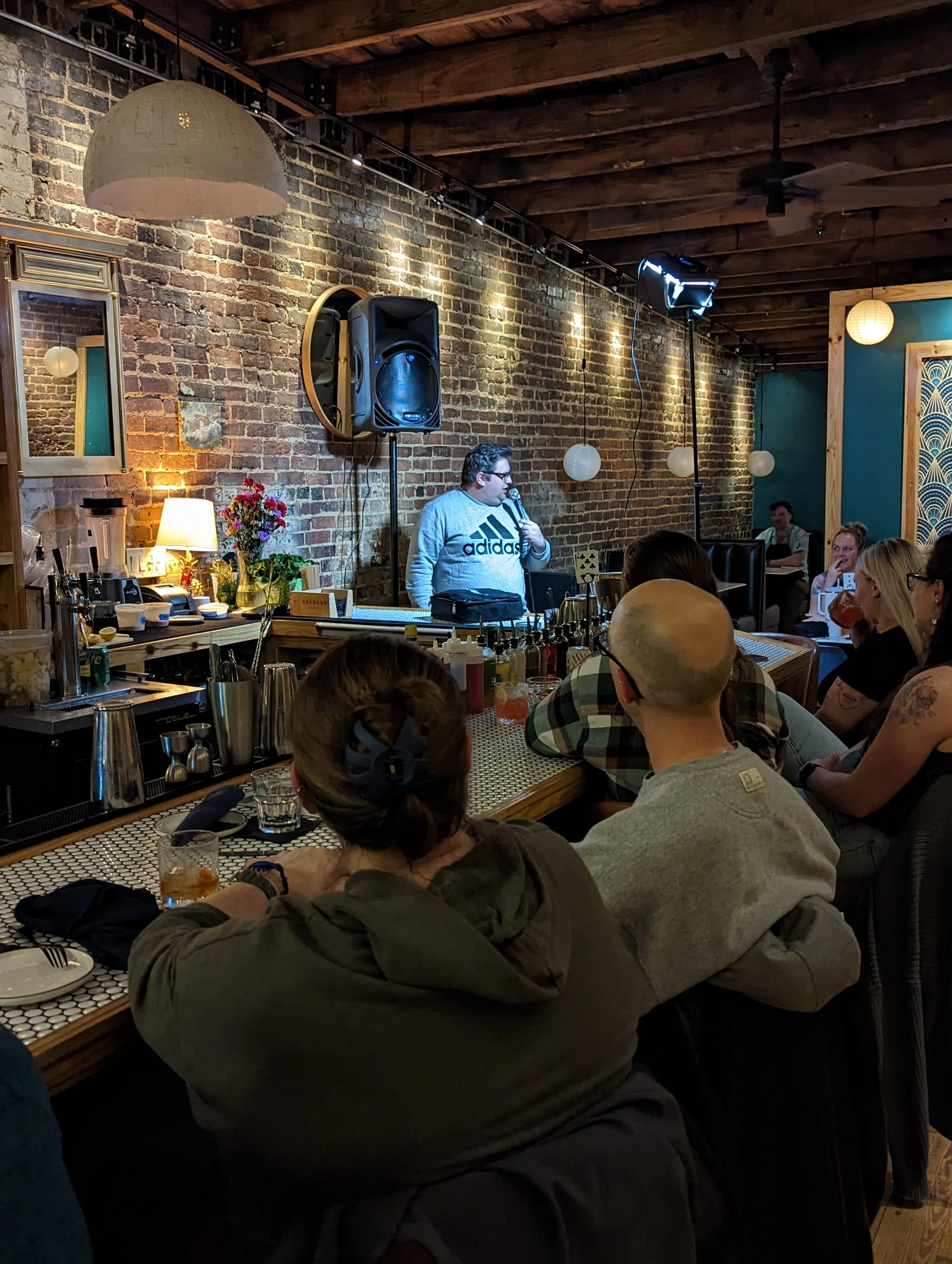 A man wearing glasses and a gray Adidas sweatshirt stands behind a bar with a microphone, performing in front of an audience in a cozy venue with exposed brick walls and wooden ceiling beams, while several audience members sit at tables enjoying drin