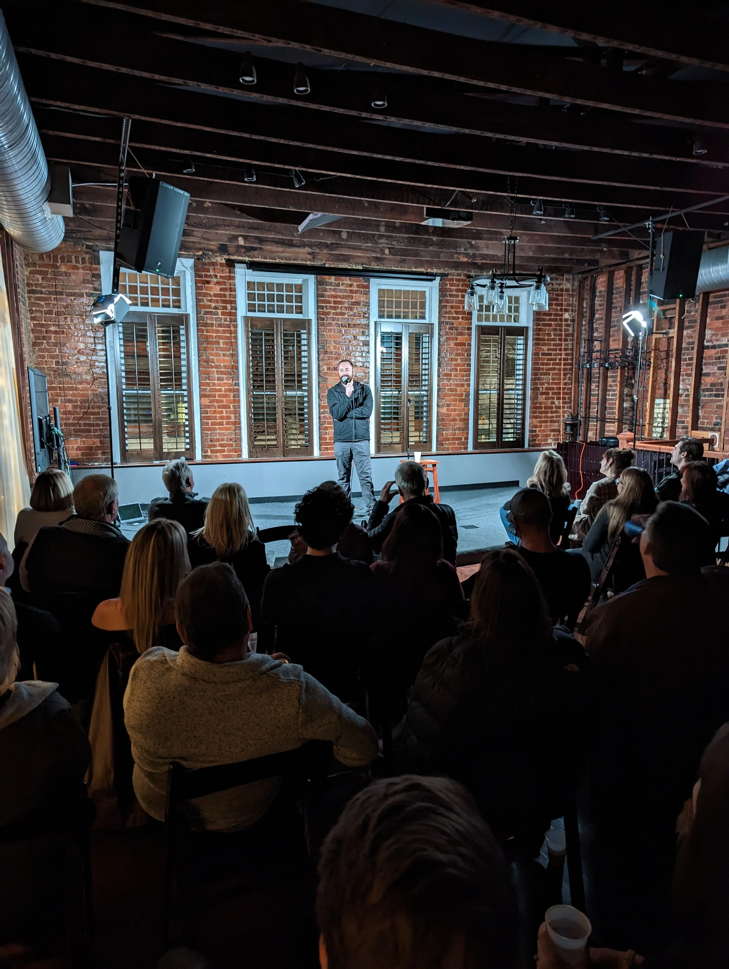 A man in a black jacket standing on a small stage, speaking or performing to a seated audience in a brick-walled venue with large windows and track lighting overhead.