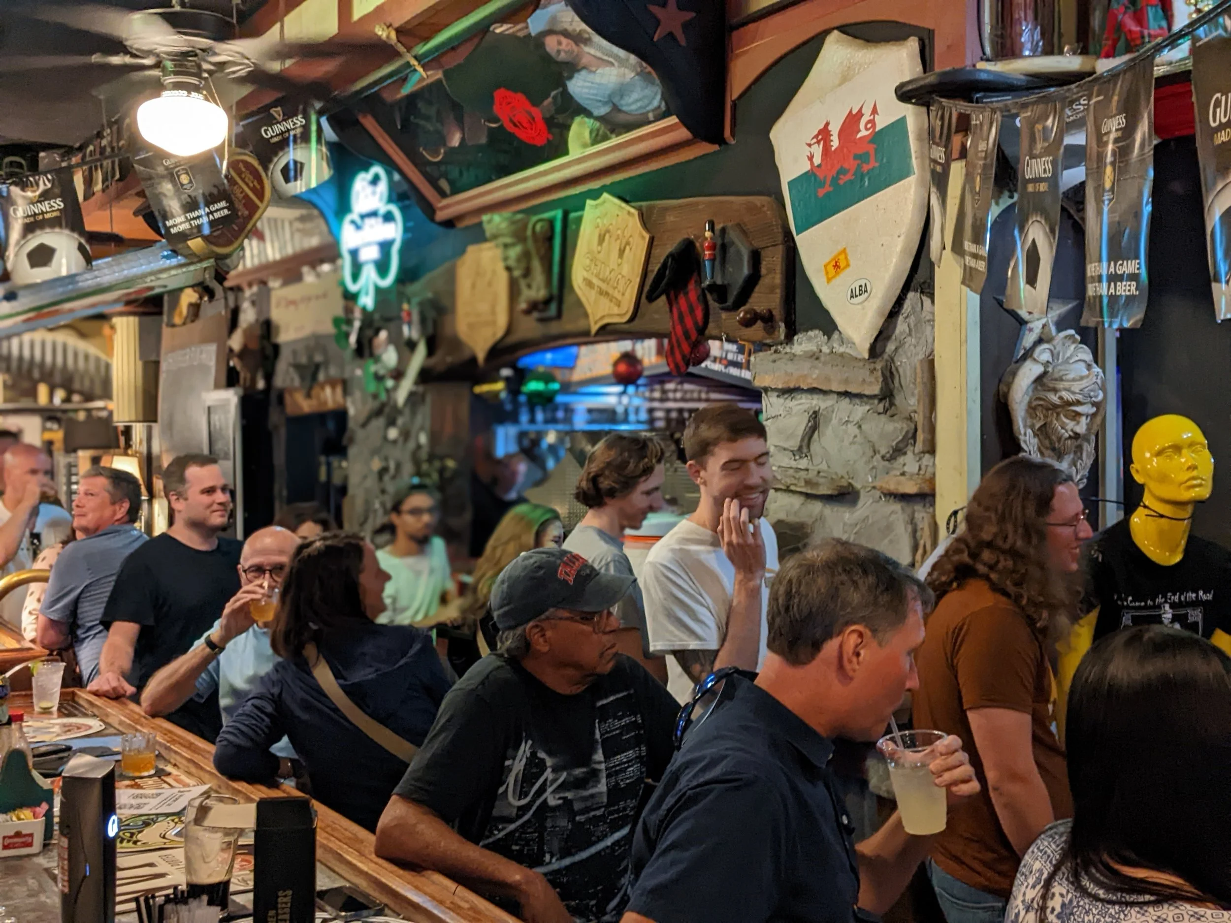 A crowded bar or pub filled with people socializing, pouring drinks, and enjoying the atmosphere. Decor includes banners, flags, and various decorations on the walls.