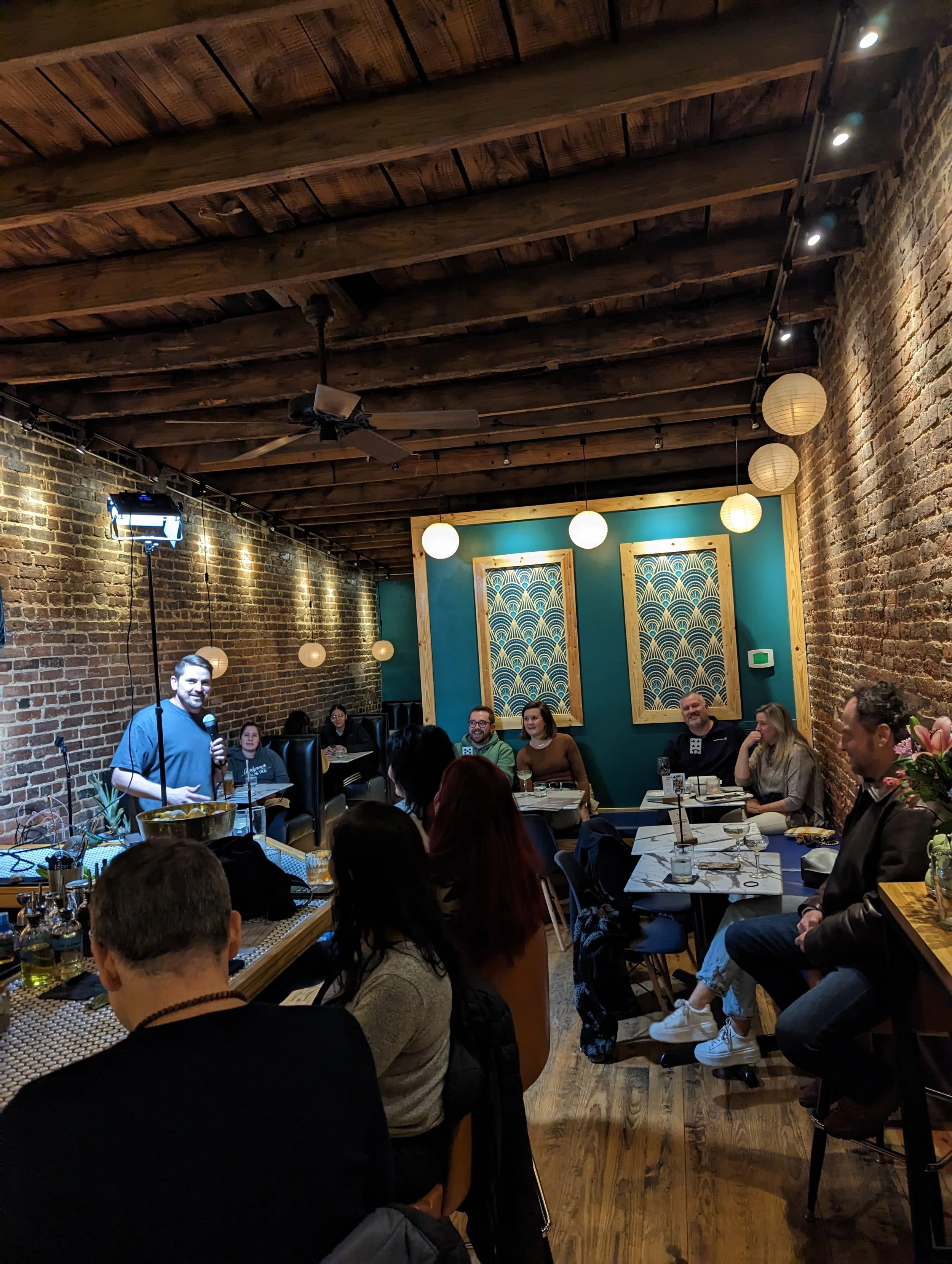 A group of people seated around tables in a restaurant with brick walls and wooden beams on the ceiling, listening to a man holding a microphone.