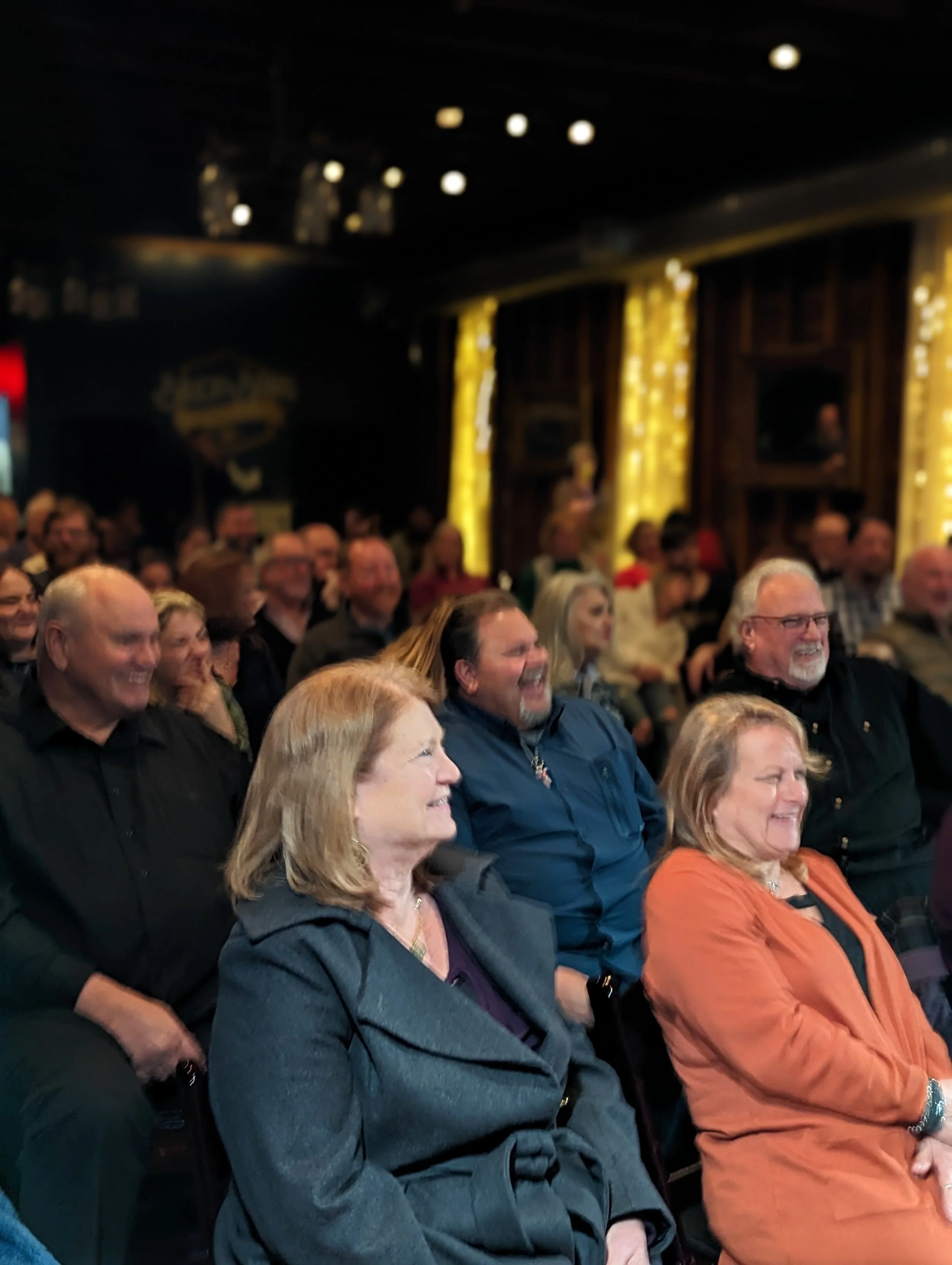 A large audience of diverse adults filled the theater, sitting in rows of chairs, watching a performance or presentation in a dimly lit indoor venue with decorative yellow curtains and dark wood paneling.