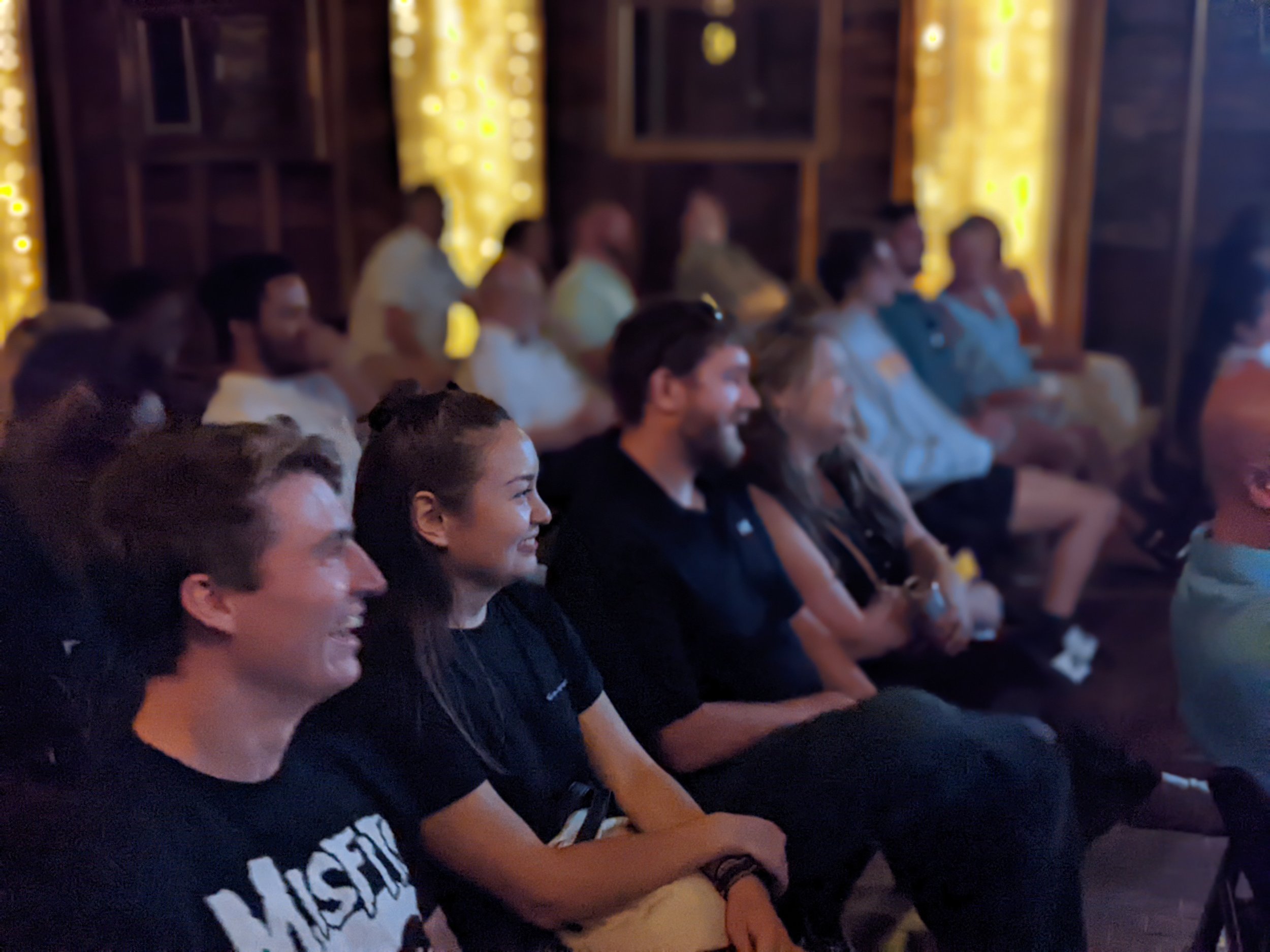 A group of people sitting in rows, watching a presentation or performance in a dimly lit room with wooden walls and decorative yellow lighting.