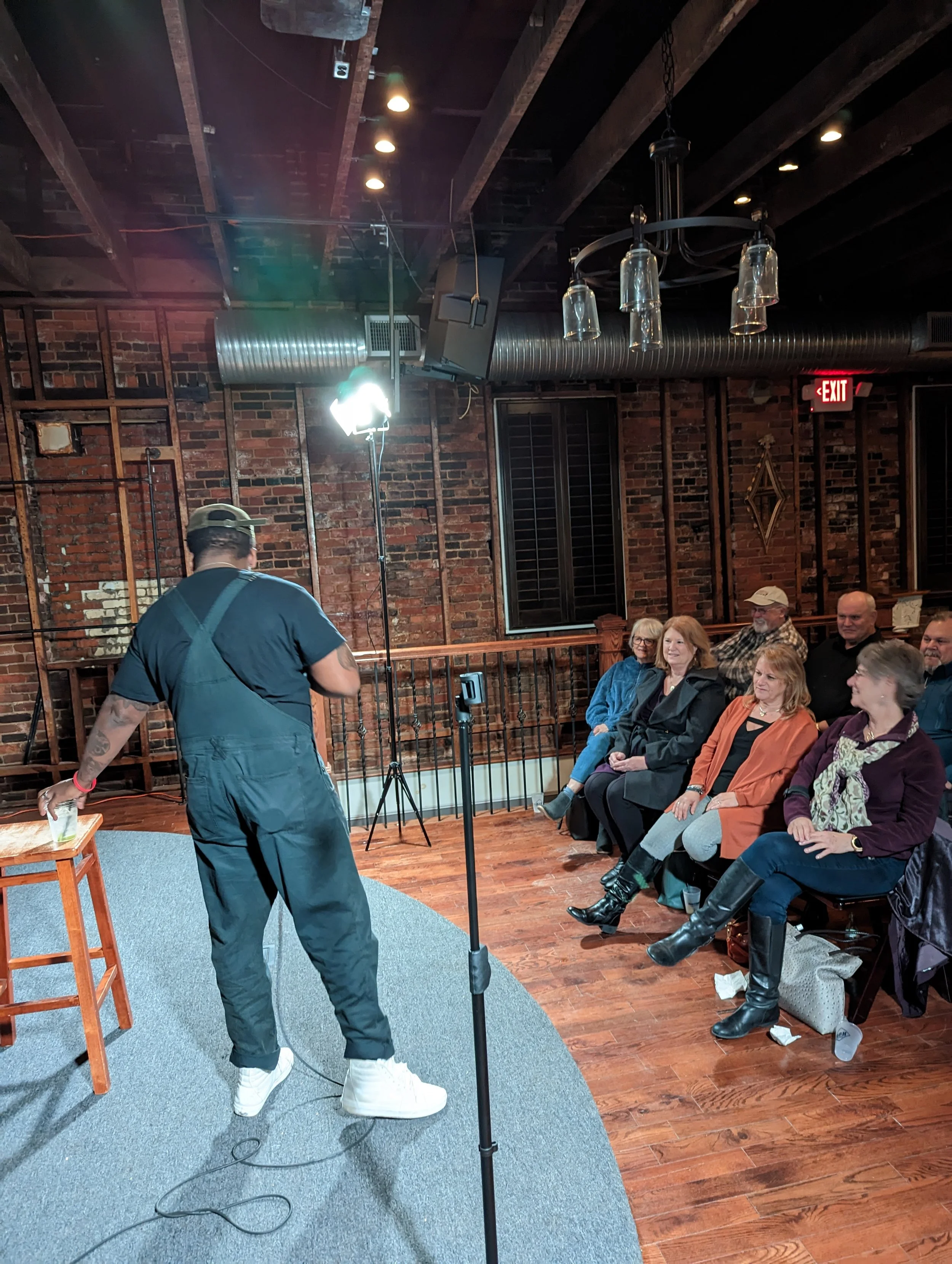 People seated in a row at an indoor event, with a man standing on a stage or platform in front of them, wearing a hat and casual clothing, with a brick wall and ceiling with exposed beams in the background.