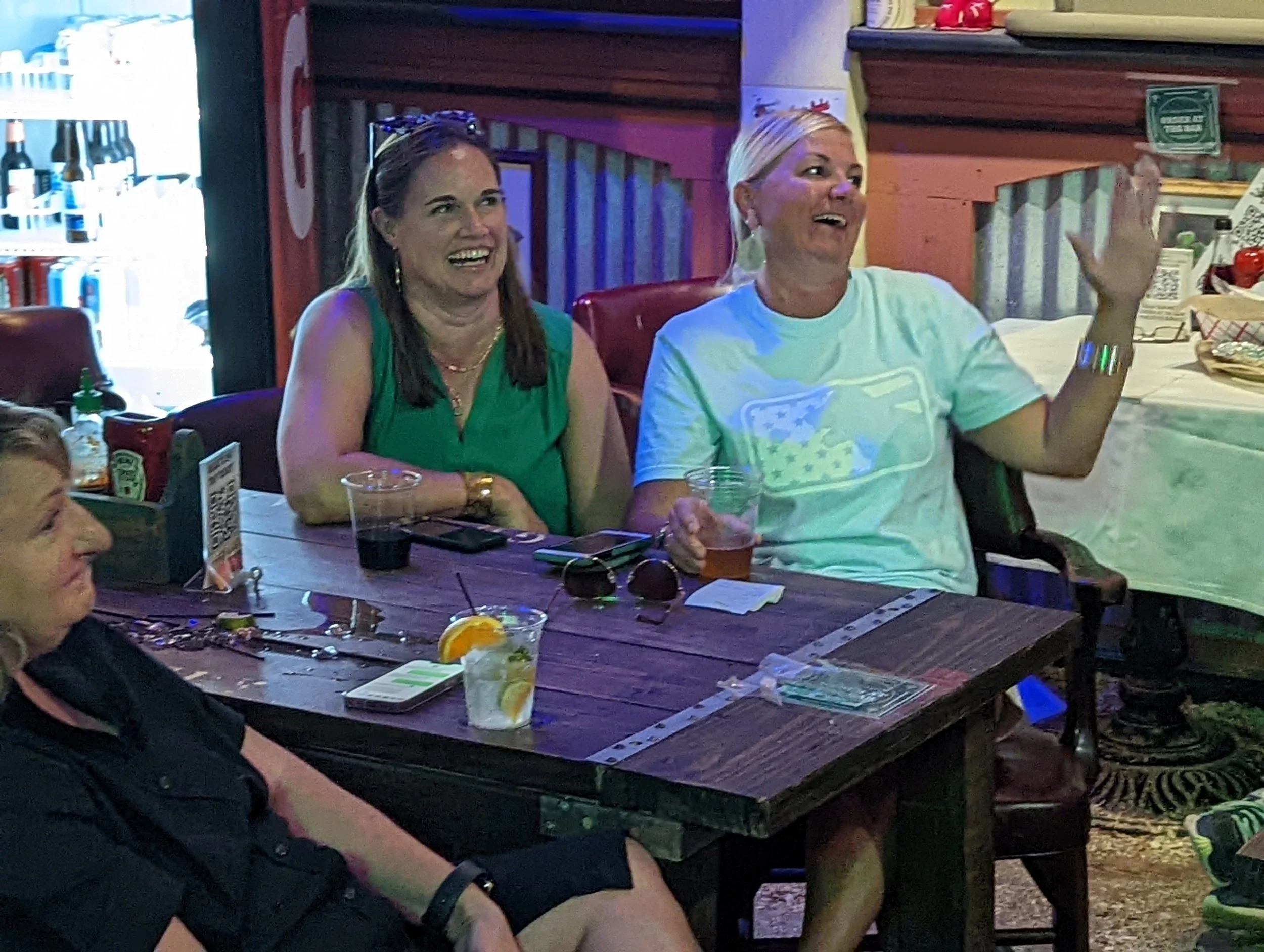 A group of people sitting at a wooden table in a casual indoor setting, appearing to enjoy a social gathering or celebration, with drinks and snacks on the table.