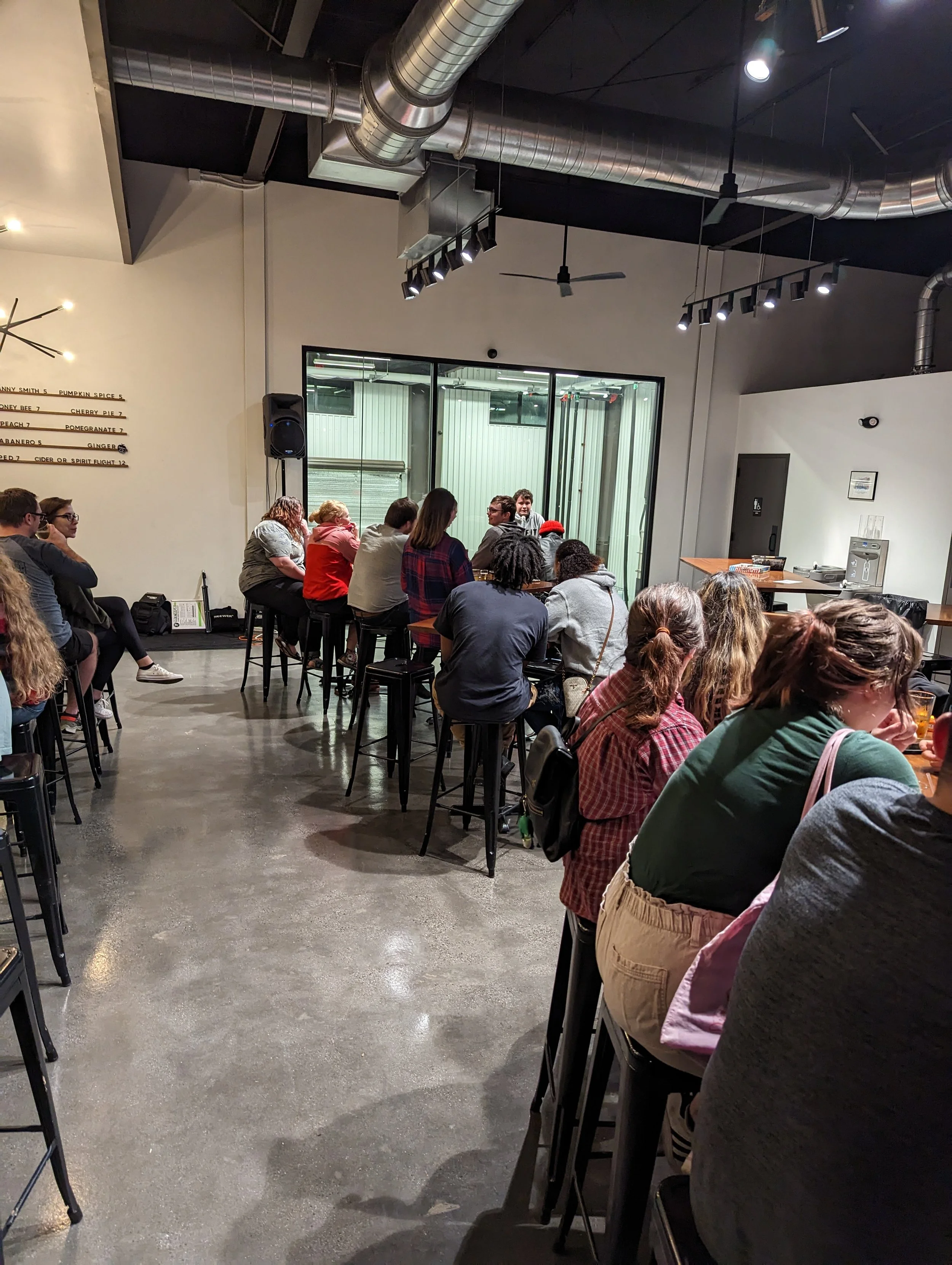 A group of people sitting on barstools inside a modern, industrial-style restaurant or brewery with high ceilings, exposed ductwork, and minimal decor.