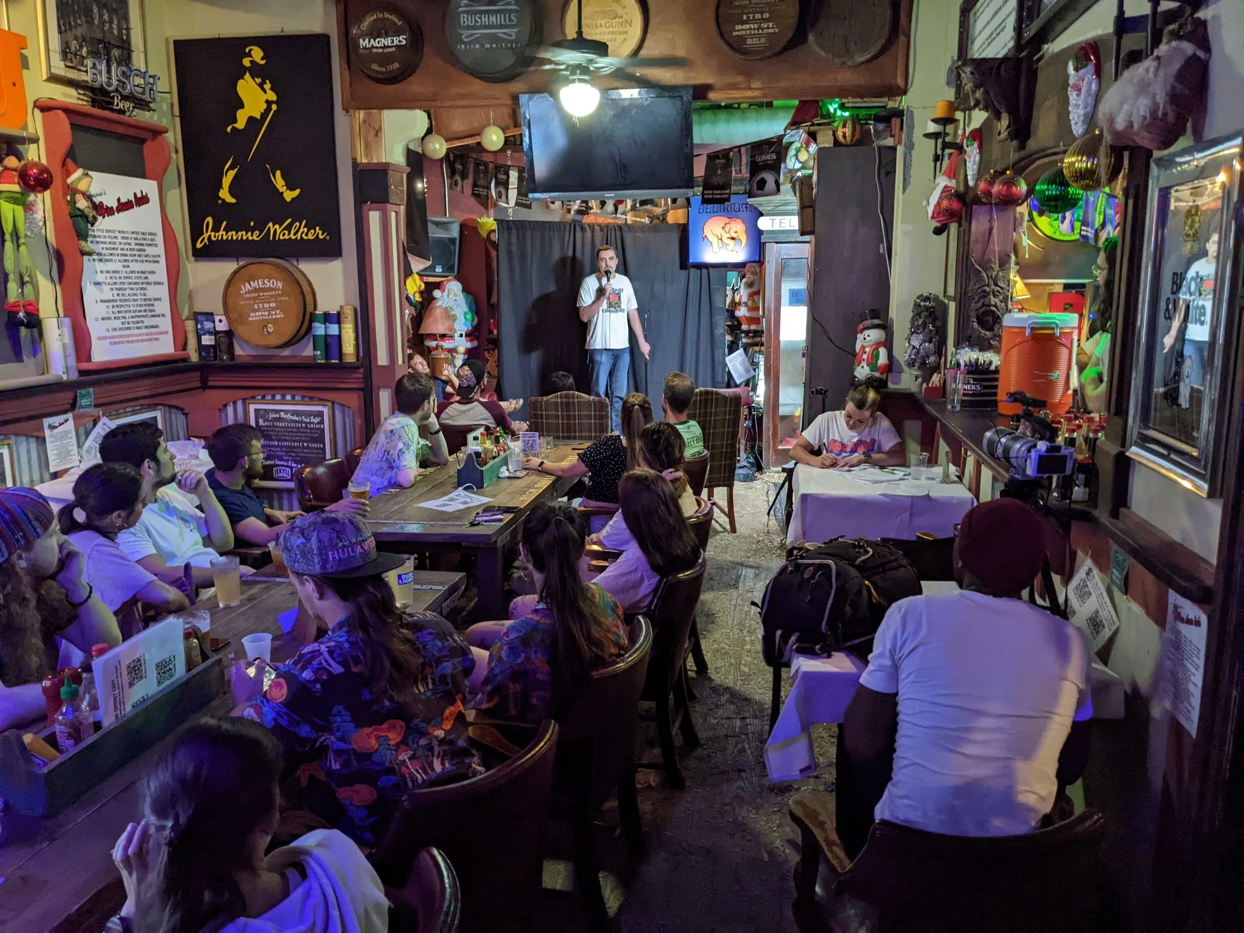 An indoor comedy show with an audience watching a performer on stage. The room has various decorations, including signs, beer barrels, and holiday ornaments.