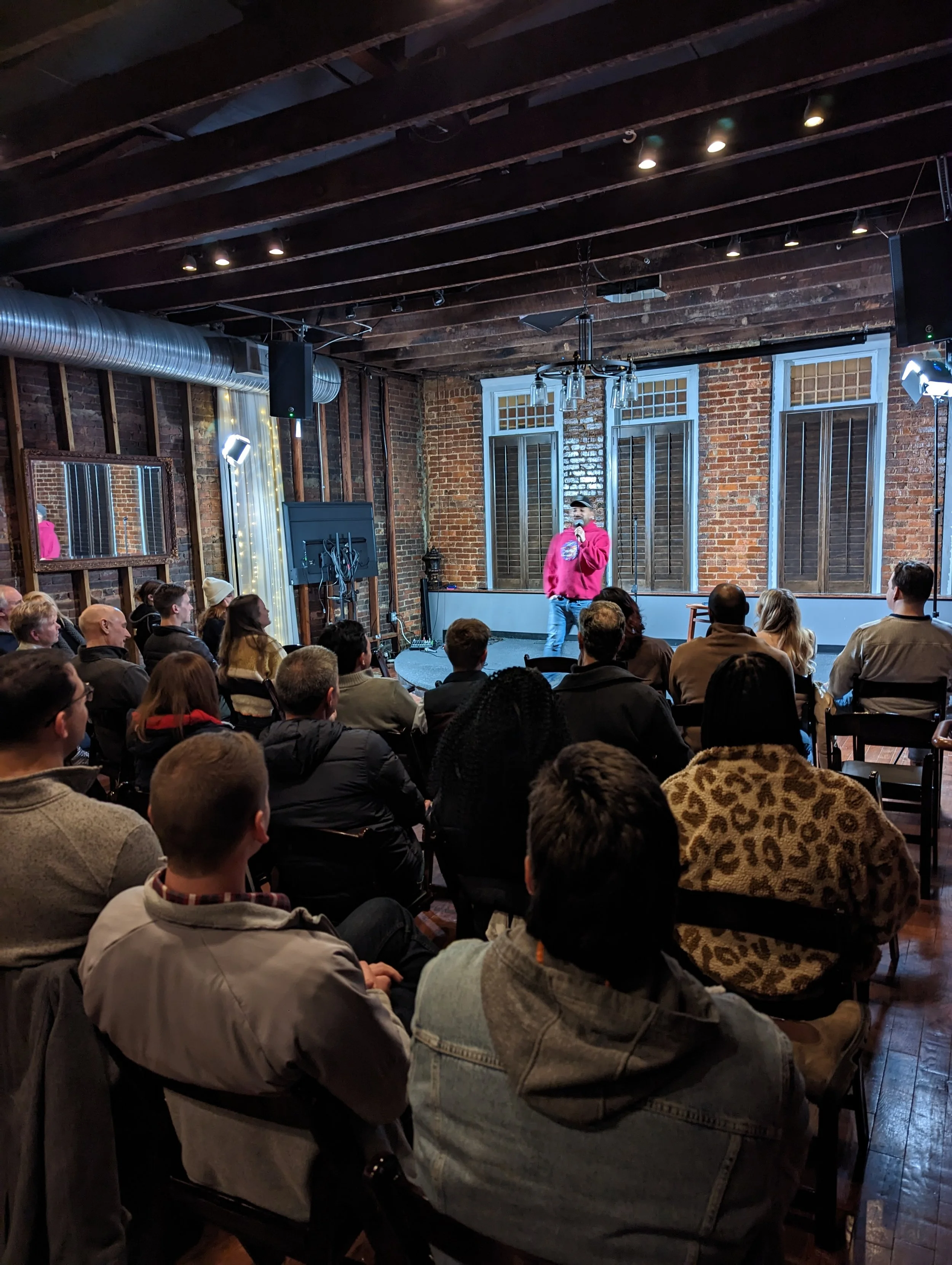 A comedian performing on stage in front of an audience in a brick-walled venue. The comedian is wearing a red hoodie and holding a microphone, with audience members seated and attentively watching.