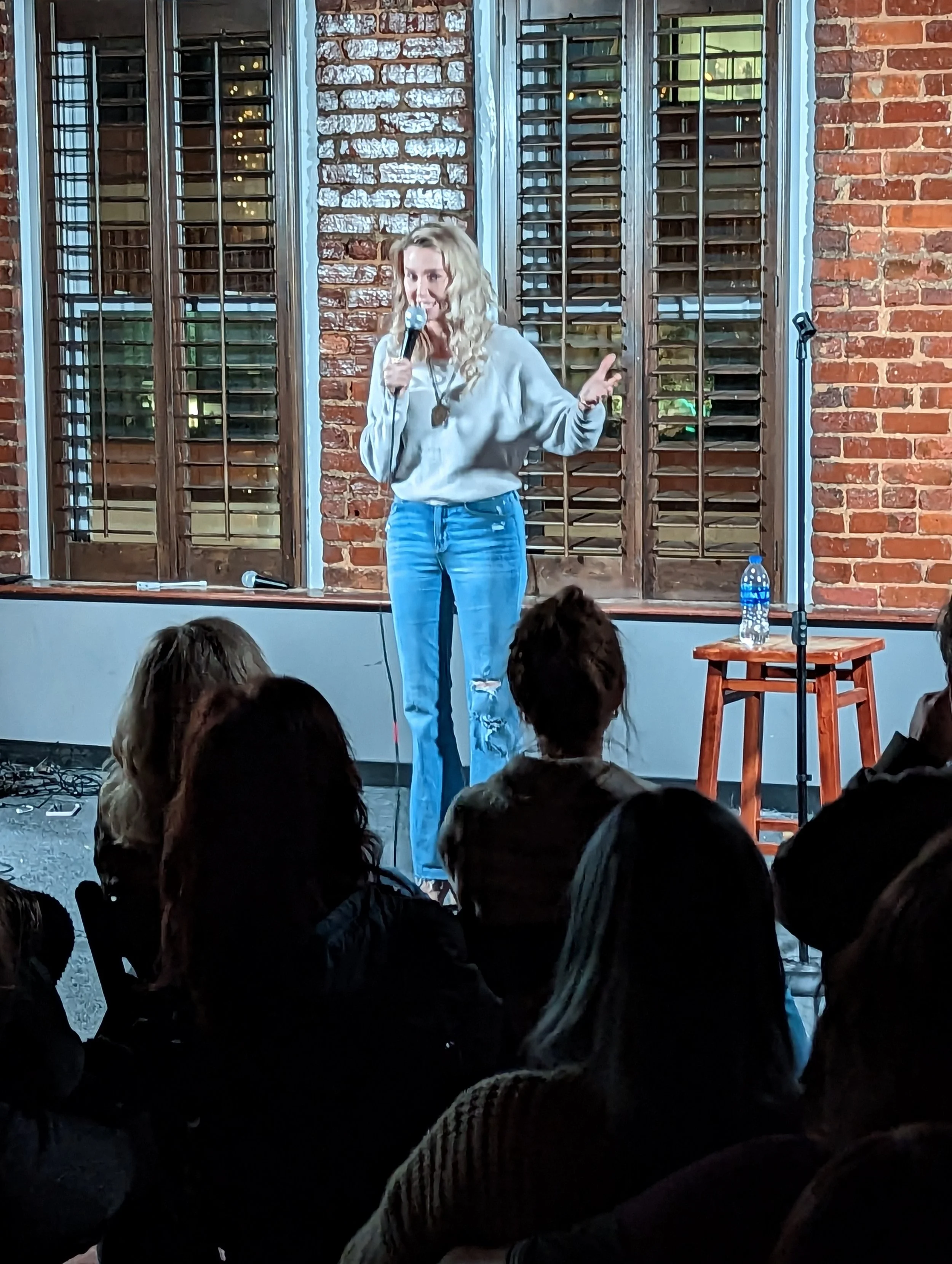 A woman with blonde curly hair stands on stage holding a microphone, speaking to an audience in a room with brick walls and wooden shutters. There is a water bottle and a microphone on a stand nearby.