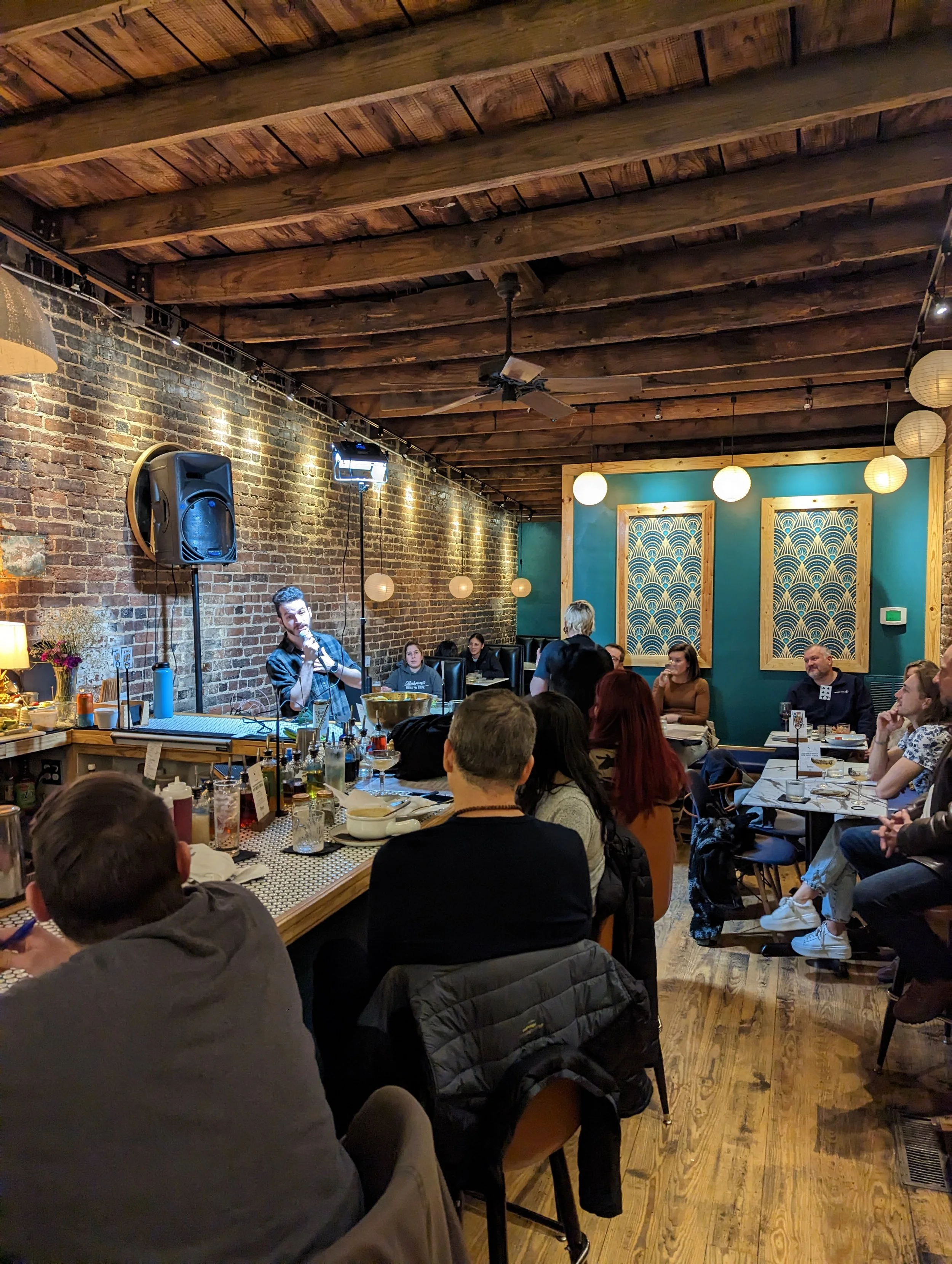 People sitting around tables and listening to a performer singing or speaking in a cozy restaurant or cafe with a brick wall, teal-colored wall, and hanging paper lanterns.
