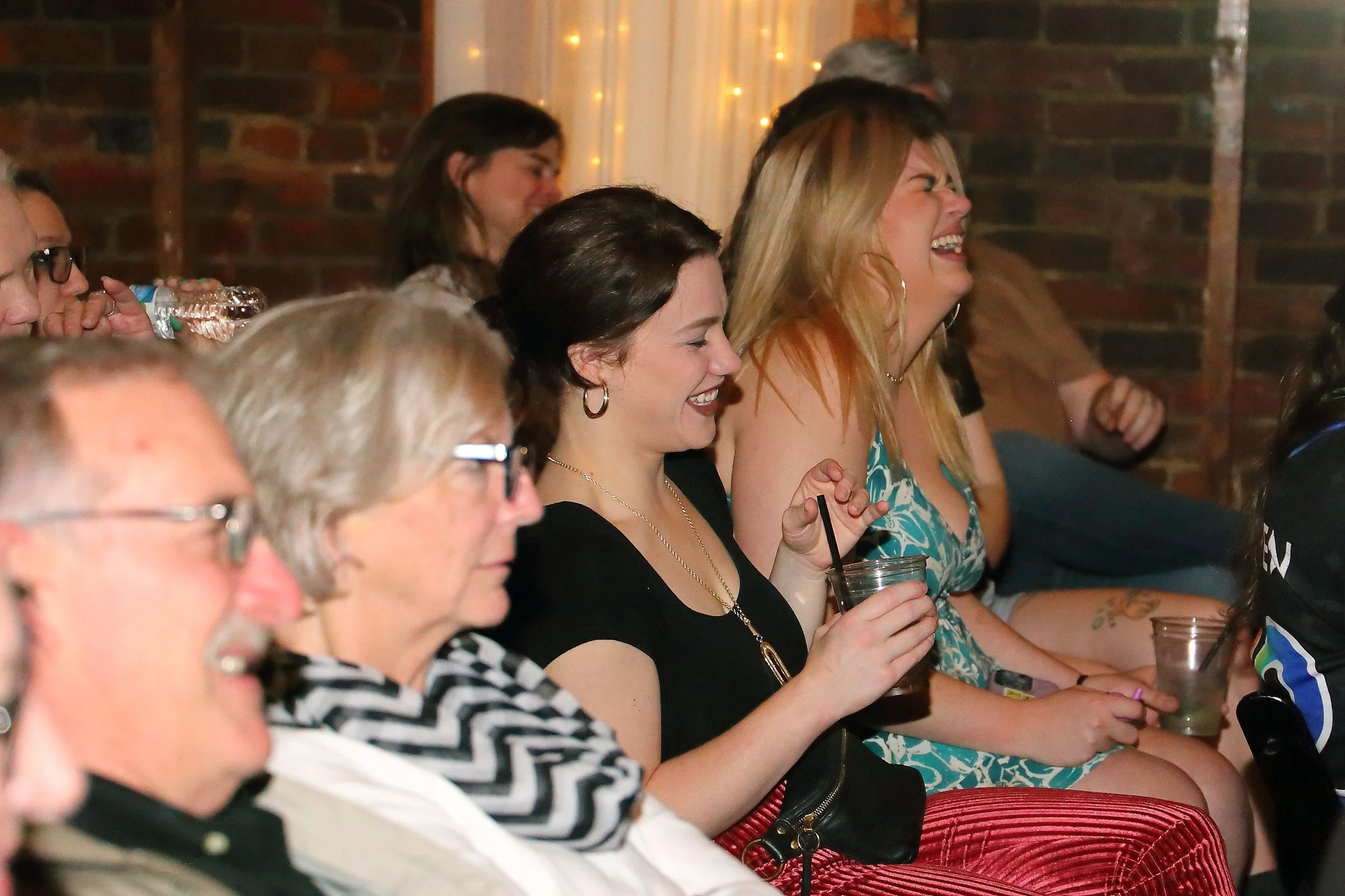 People sitting in a row at a social gathering, laughing and enjoying drinks, with a brick wall and string lights in the background.