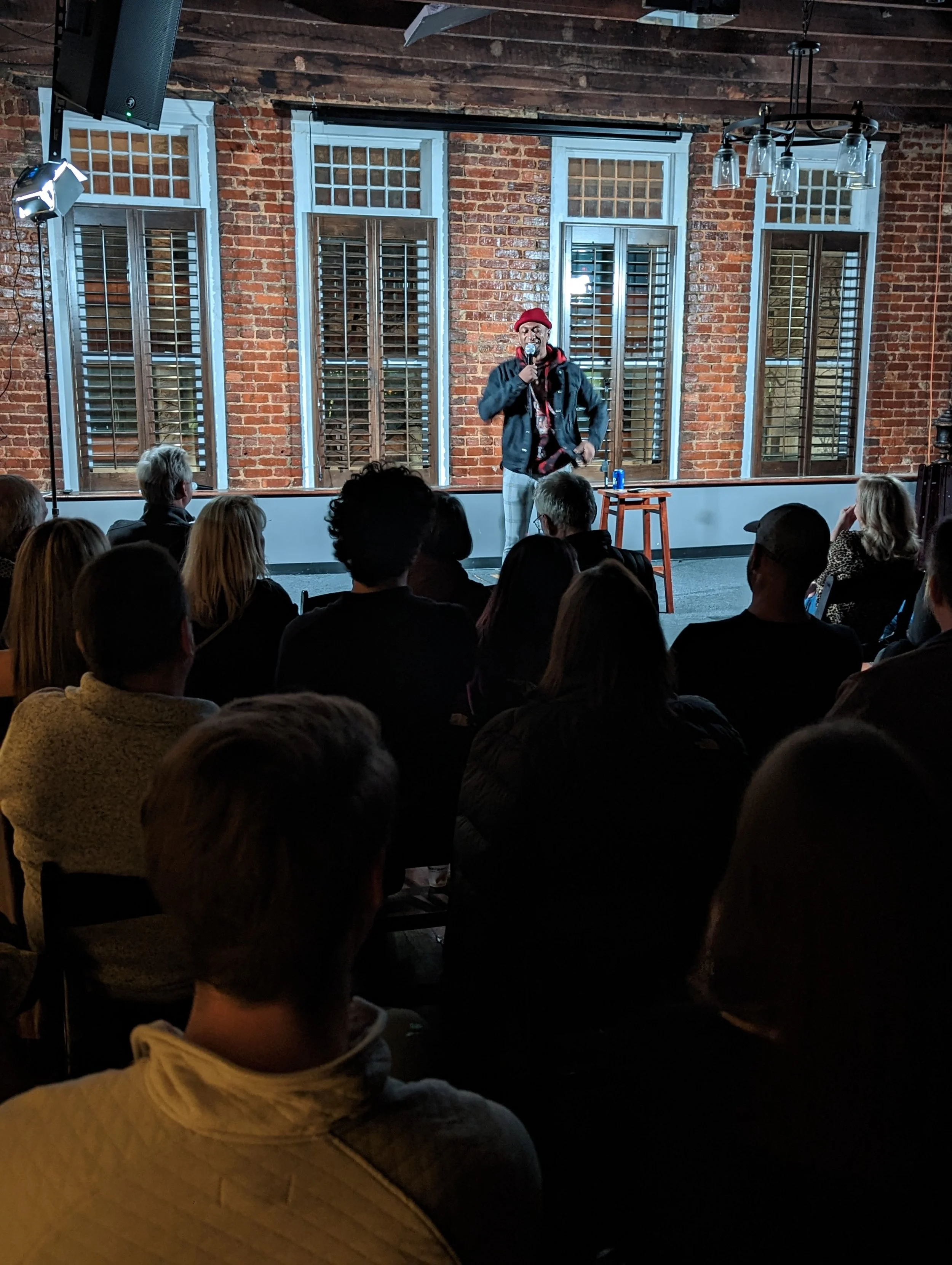 A comedian stands on stage holding a microphone, wearing a red beanie, a black jacket, and plaid pants, performing in front of an audience in a venue with exposed brick walls and large windows with shutters.