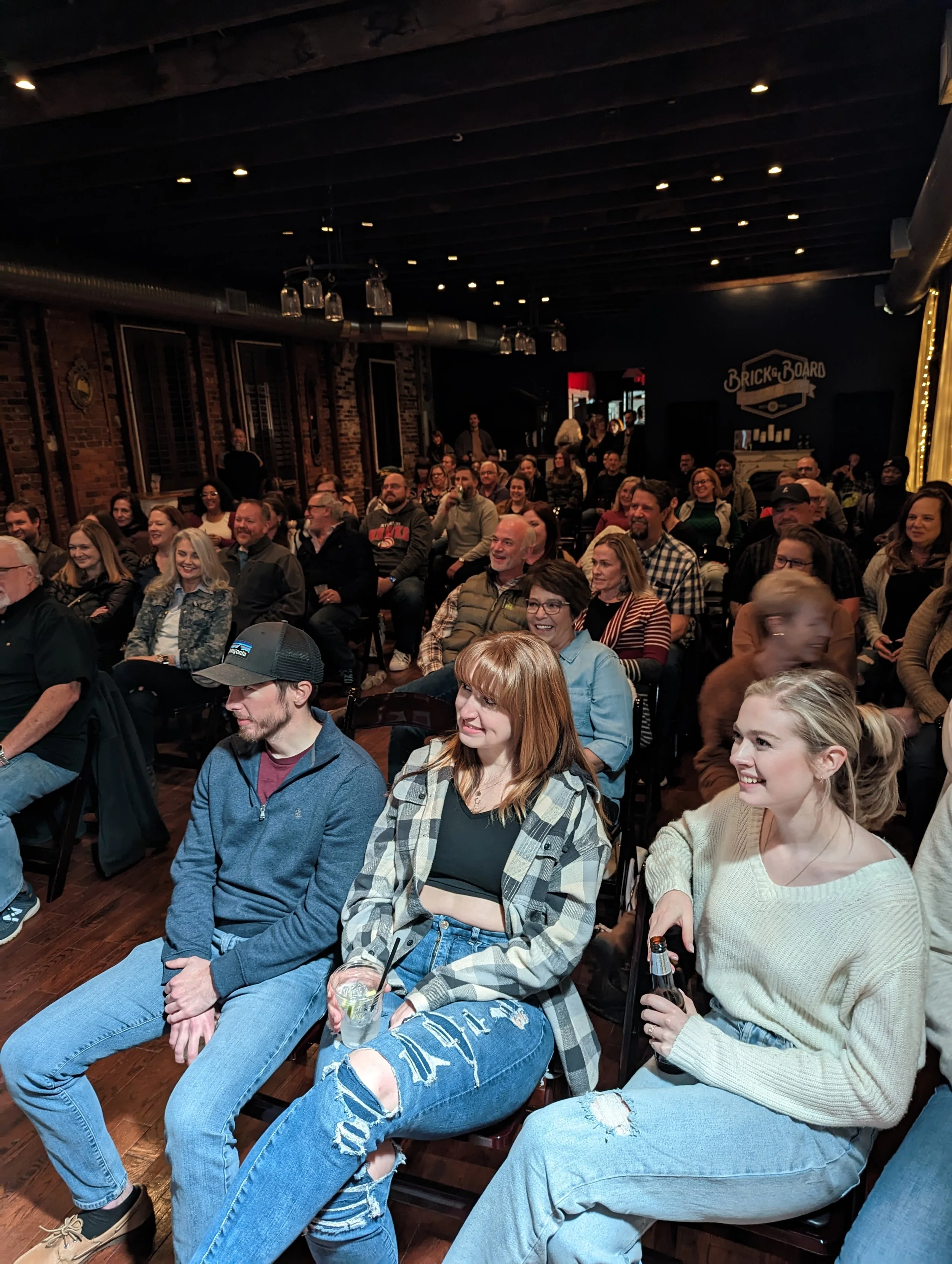 A group of people sitting in a dark, rustic venue, laughing and enjoying a live comedy show or performance.