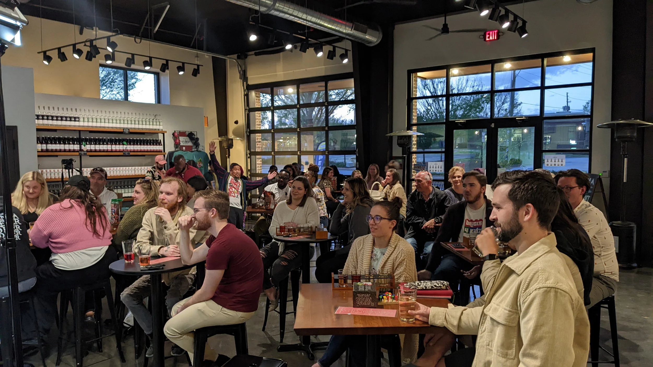 A crowded indoor restaurant or bar with people seated at tables, some enjoying drinks and conversation. The setting has large windows, industrial-style ceiling with exposed ductwork, and a shelf with bottles on the wall. The atmosphere appears lively