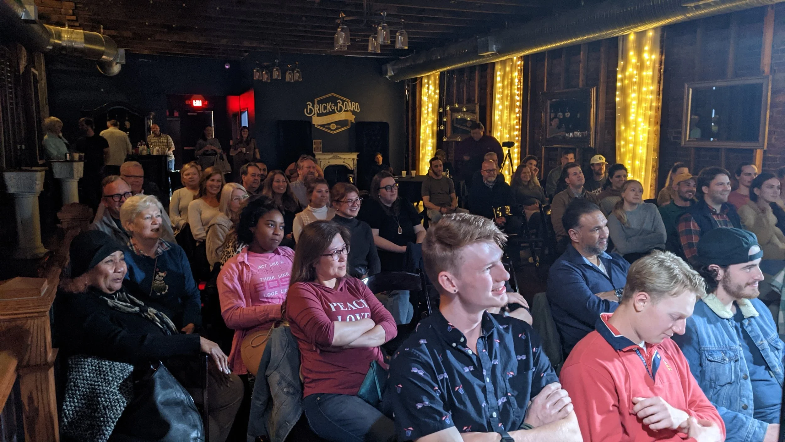 An indoor comedy show or presentation with an audience seated and smiling, decorated with string lights on the walls.