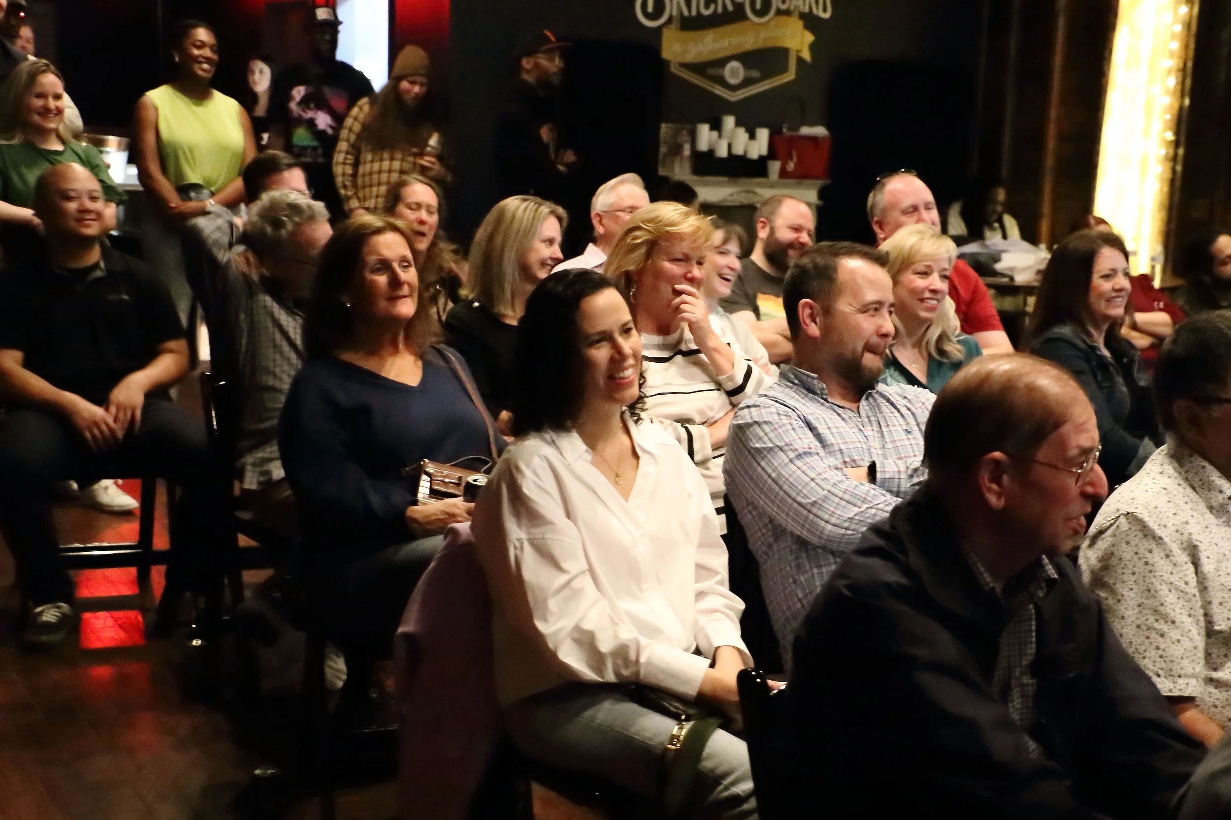 A group of people seated and laughing in an indoor venue, likely watching a performance or speaker, with a few standing in the background, some taking photos.