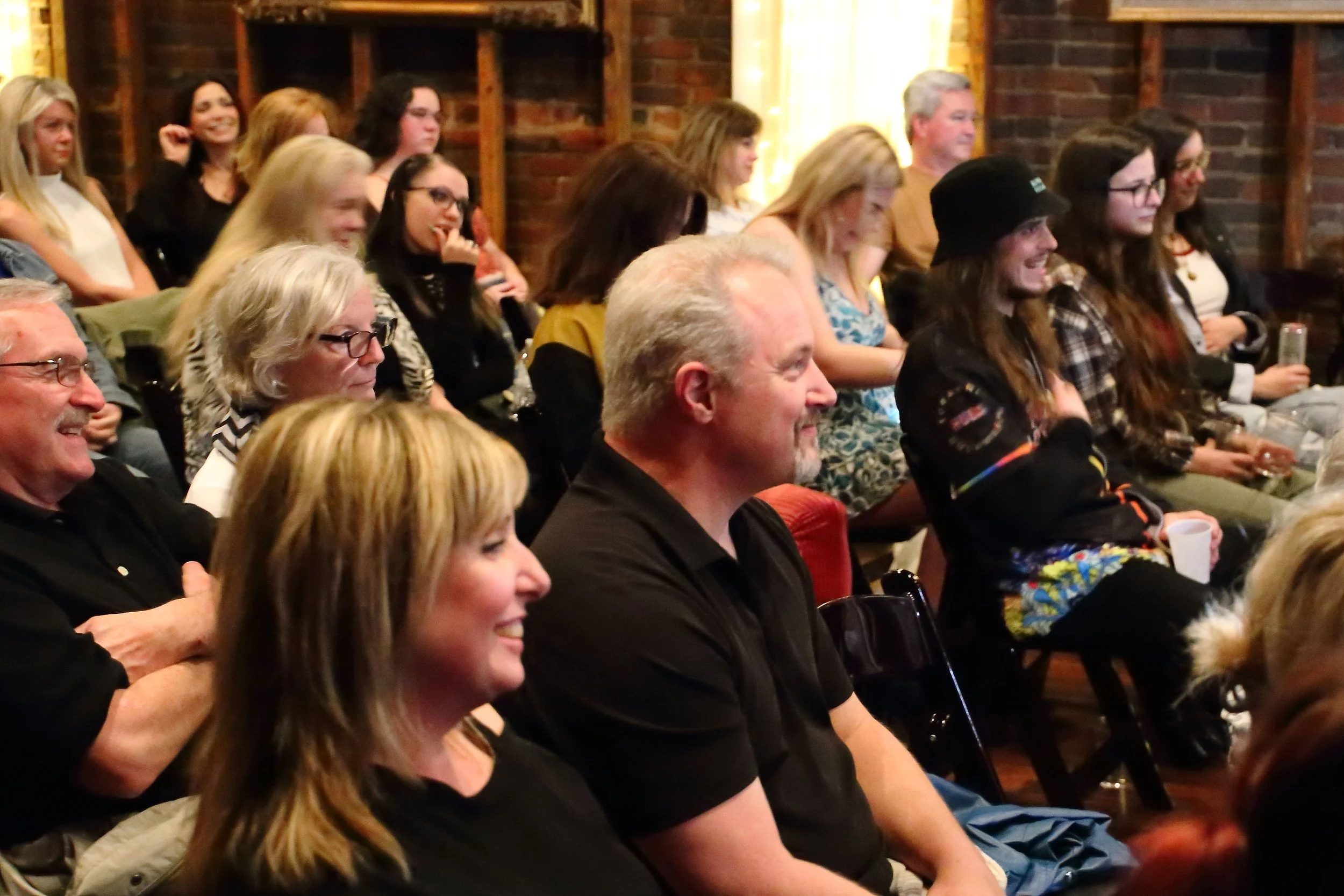 Audience members sitting and smiling during a performance or event in a rustic room with brick walls.