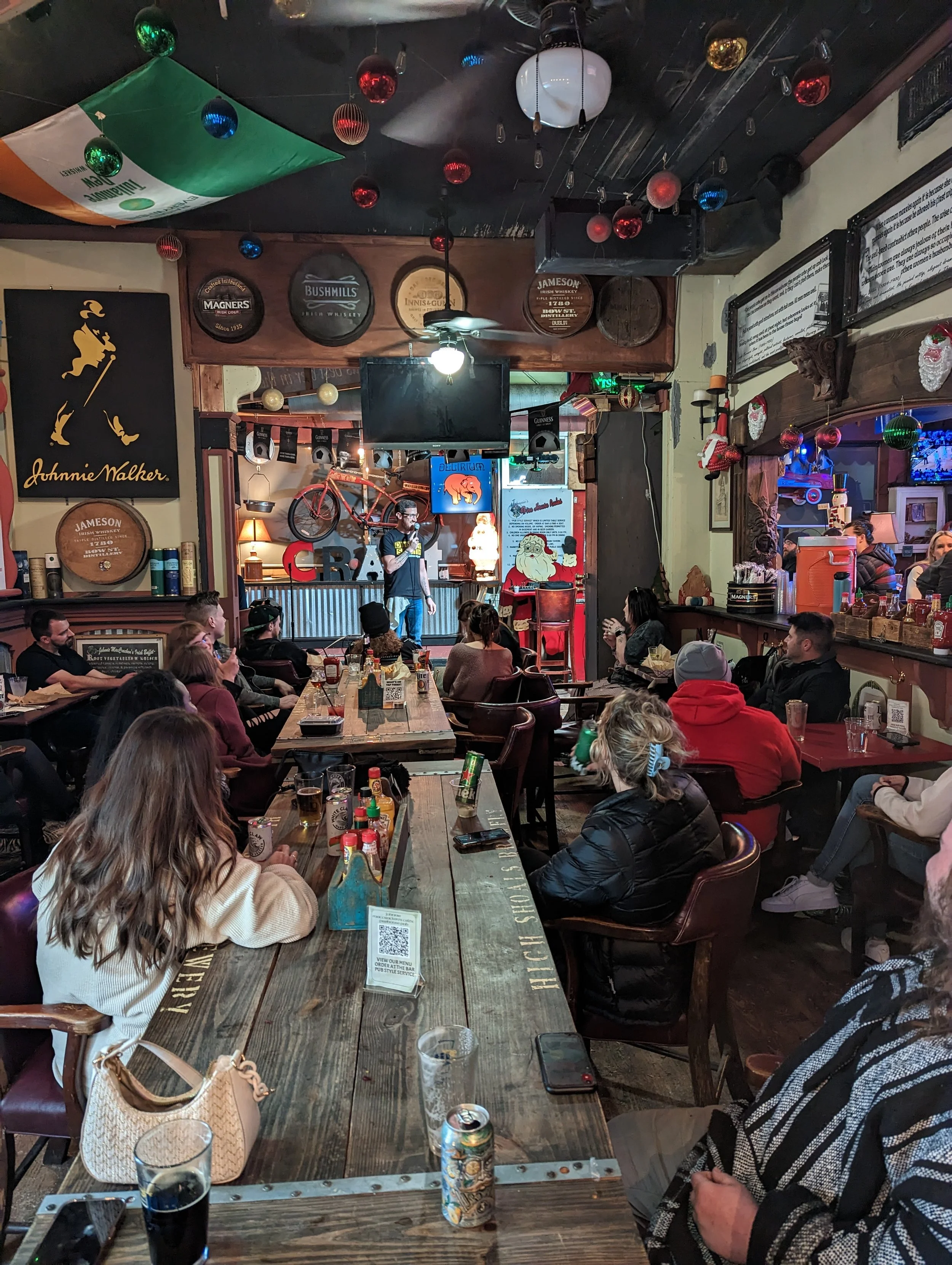People sitting and standing inside a lively bar or pub with Christmas decorations, vintage signs, and a performer on stage.