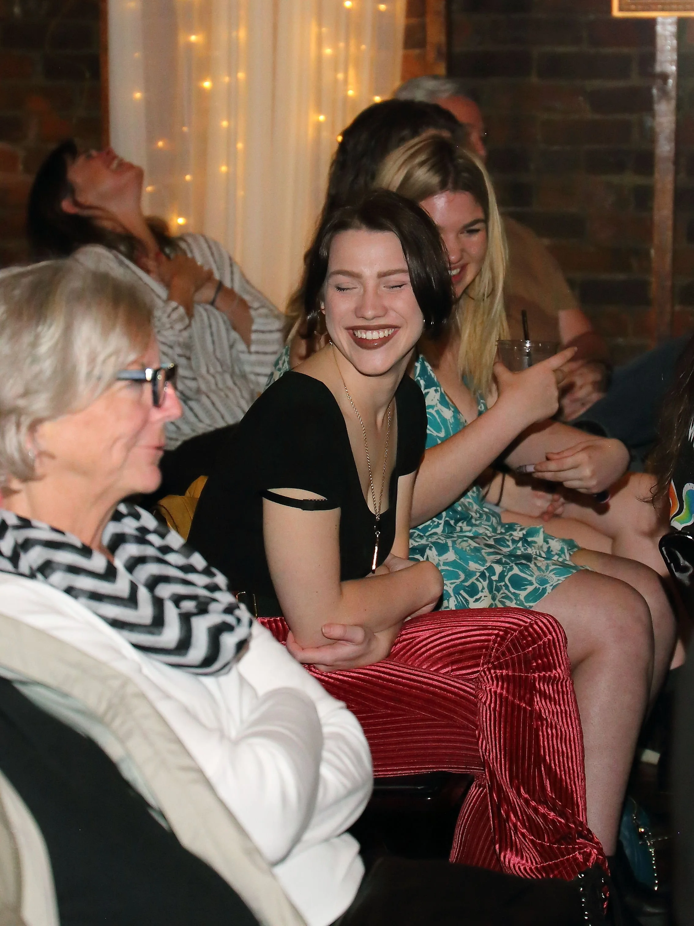 A group of people sitting in a row at a social gathering, with some smiling and others appearing to be laughing, in a warmly lit indoor setting with string lights and brick walls.
