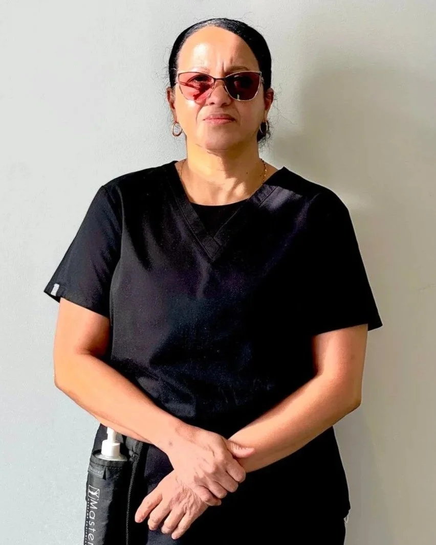 A woman with black hair, wearing sunglasses and black scrubs, standing against a light-colored wall.