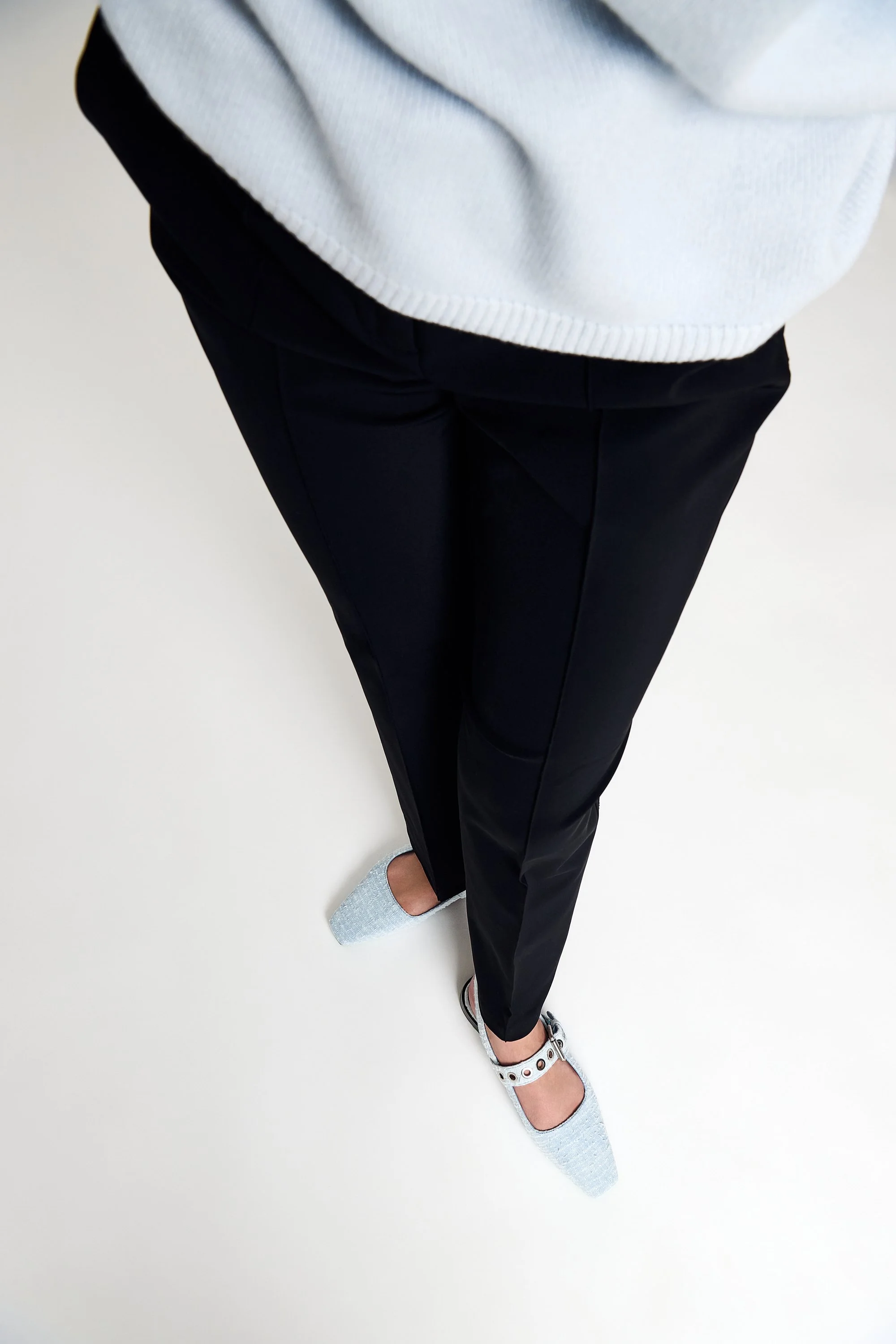 A top view photo of a person legs standing on a plain white surface, wearing black pants, a light-colored sweater, and light gray shoes with small cutouts and a strap.