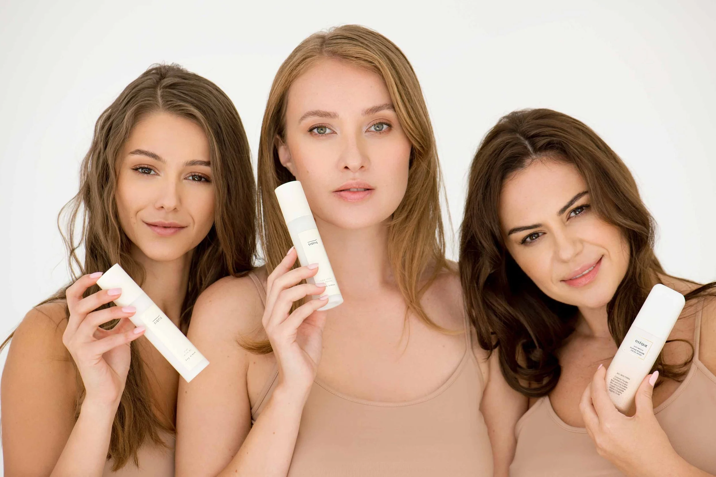 Three women with long hair holding skincare products, standing against a plain white background.