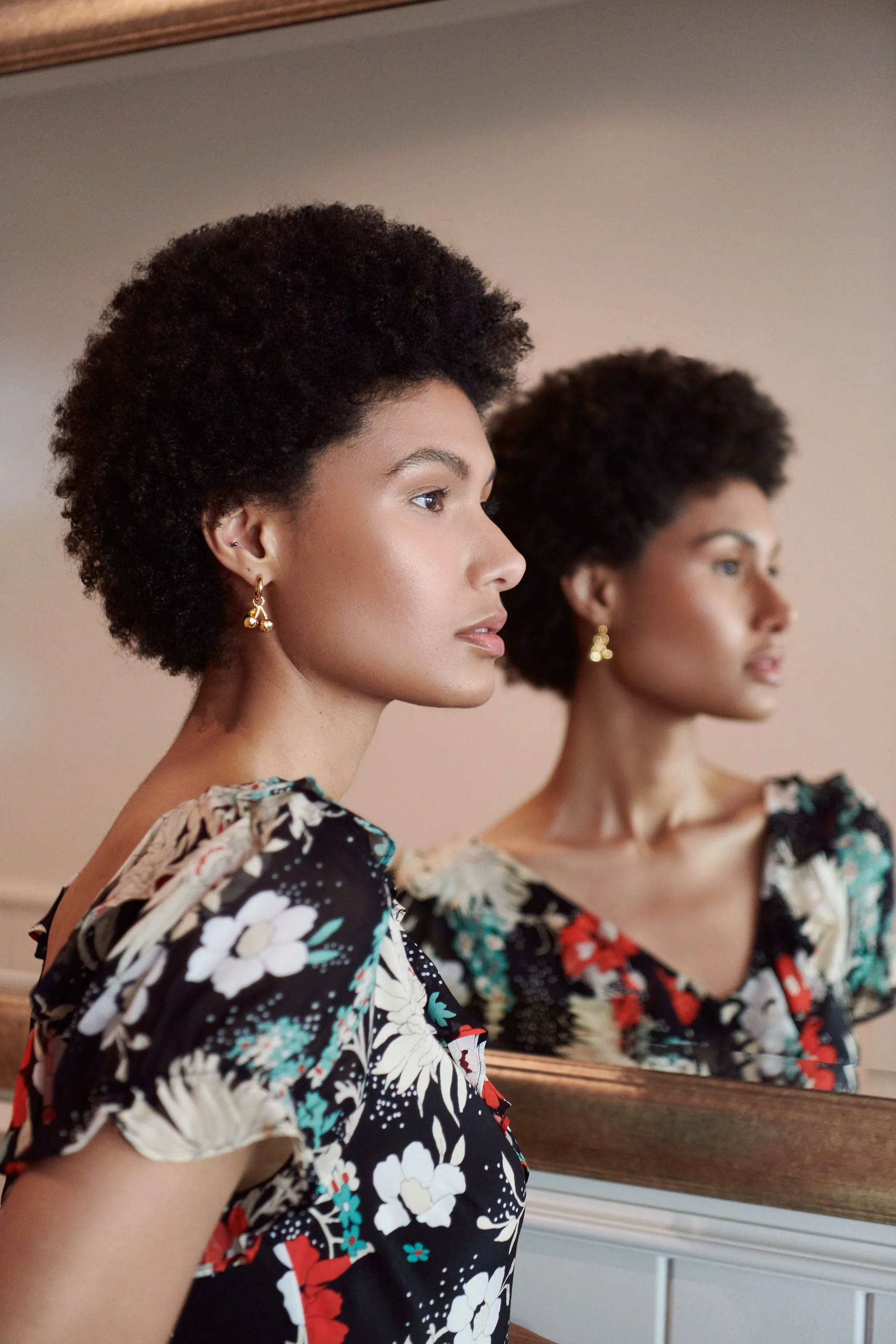 A woman with short curly hair stands in front of a mirror, dressed in a floral patterned top with black, white, red, and green colors. She is wearing gold earrings and gazes to the left, with her reflection visible in the mirror behind her.