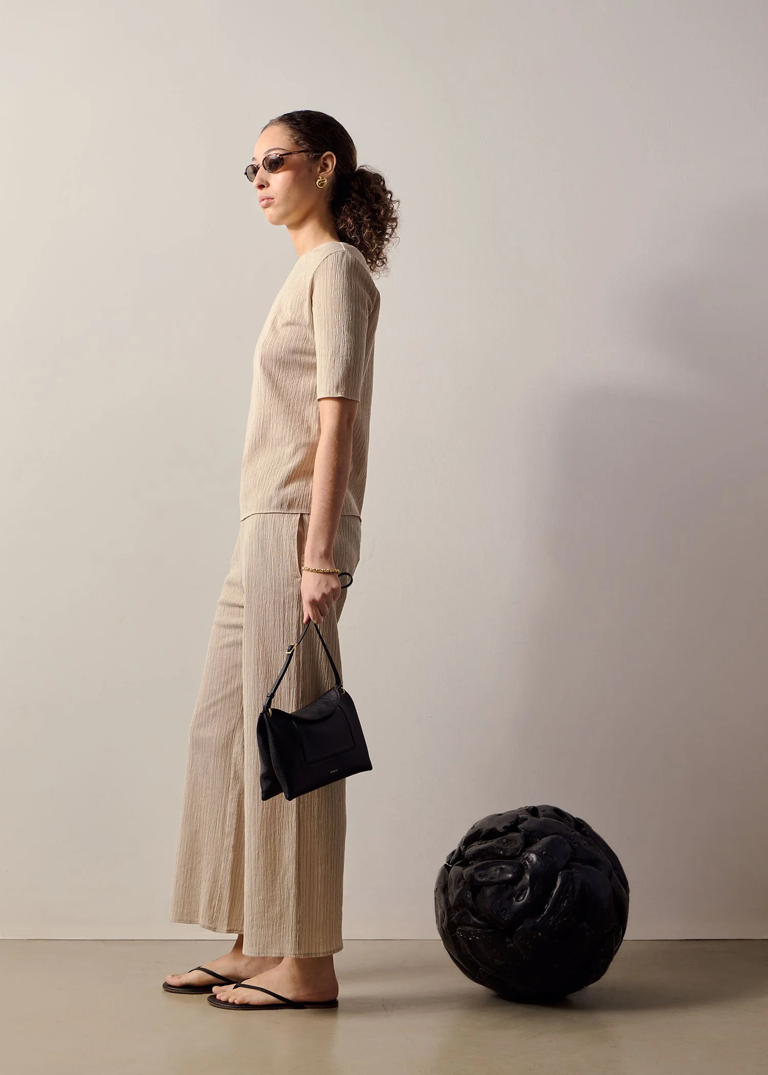 A woman in beige outfit standing in profile, holding a black handbag, on a beige floor next to a large, textured black sculpture.