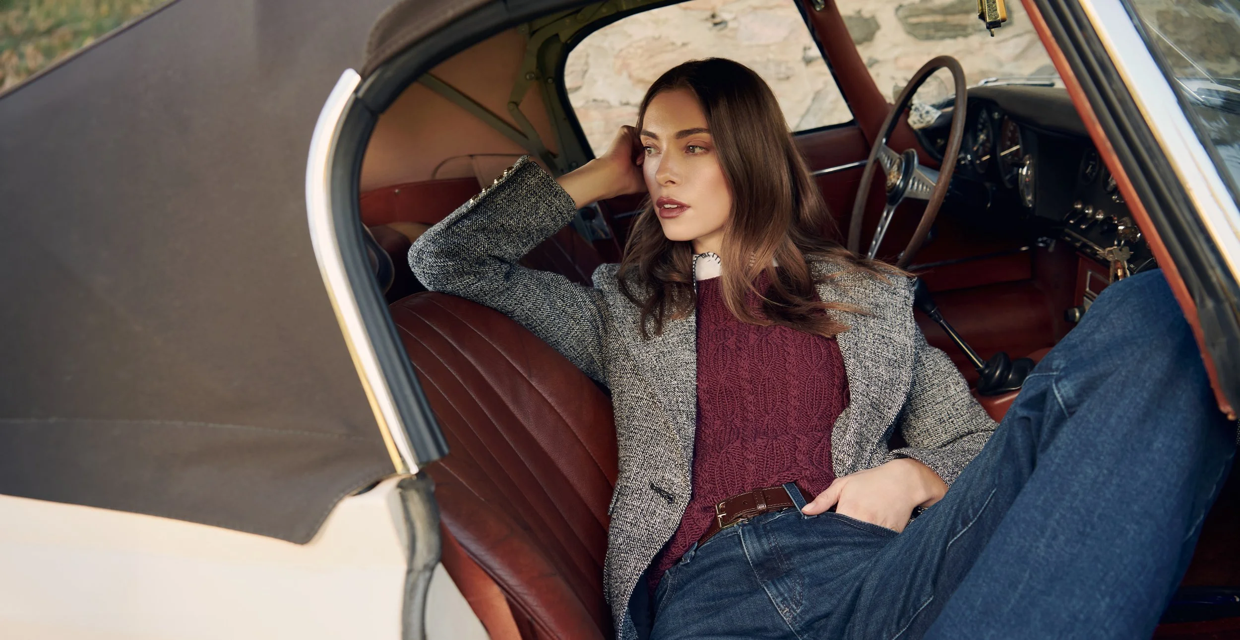 A woman lounging inside a vintage car, wearing a gray blazer, red sweater, and blue jeans, with her hand resting on her head and looking thoughtfully.