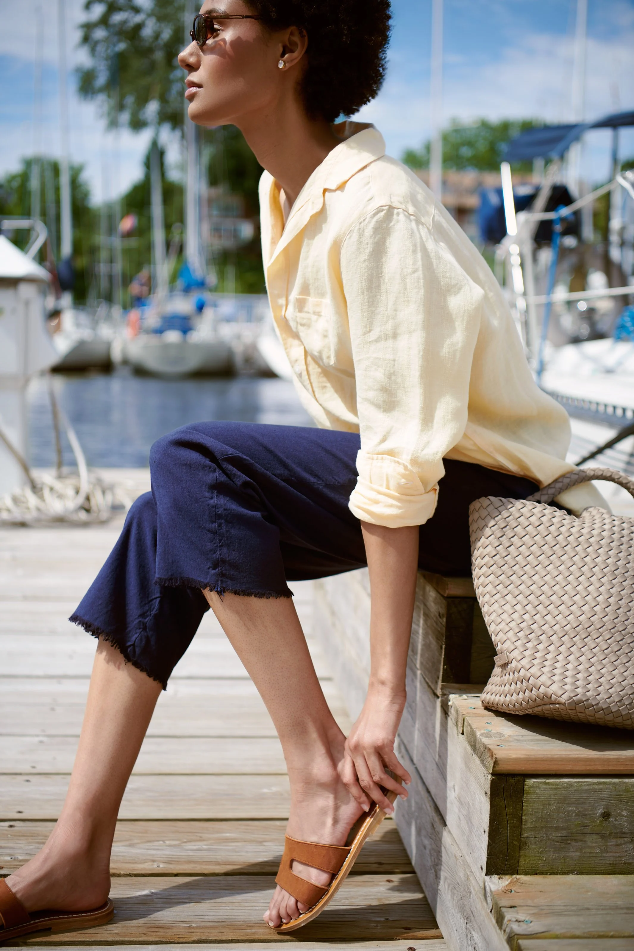 A woman with short curly hair wearing sunglasses, a light yellow shirt, and dark rolled-up jeans, is sitting on a wooden dock by a marina with boats and water in the background, adjusting her sandal.