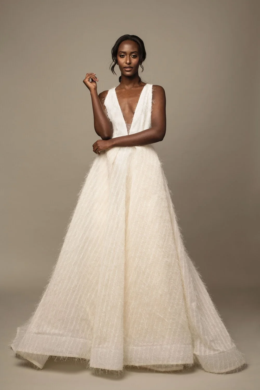 A woman wearing an elegant, cream-colored, sleeveless gown with a deep V neckline and textured fabric, standing against a plain background.