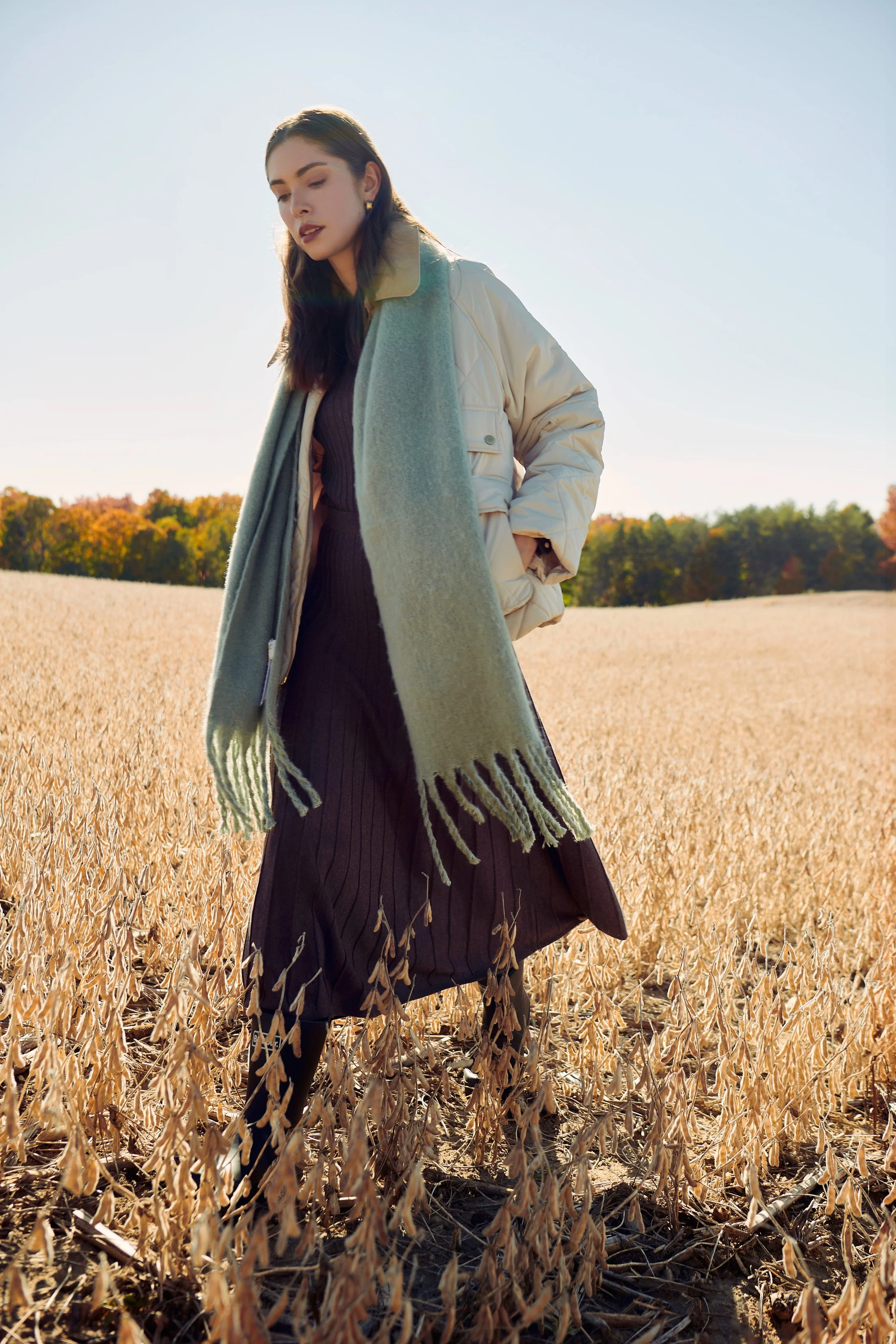 A woman with dark hair stands in a field of drying crops during daytime, wearing a beige puffer jacket and a long scarf.