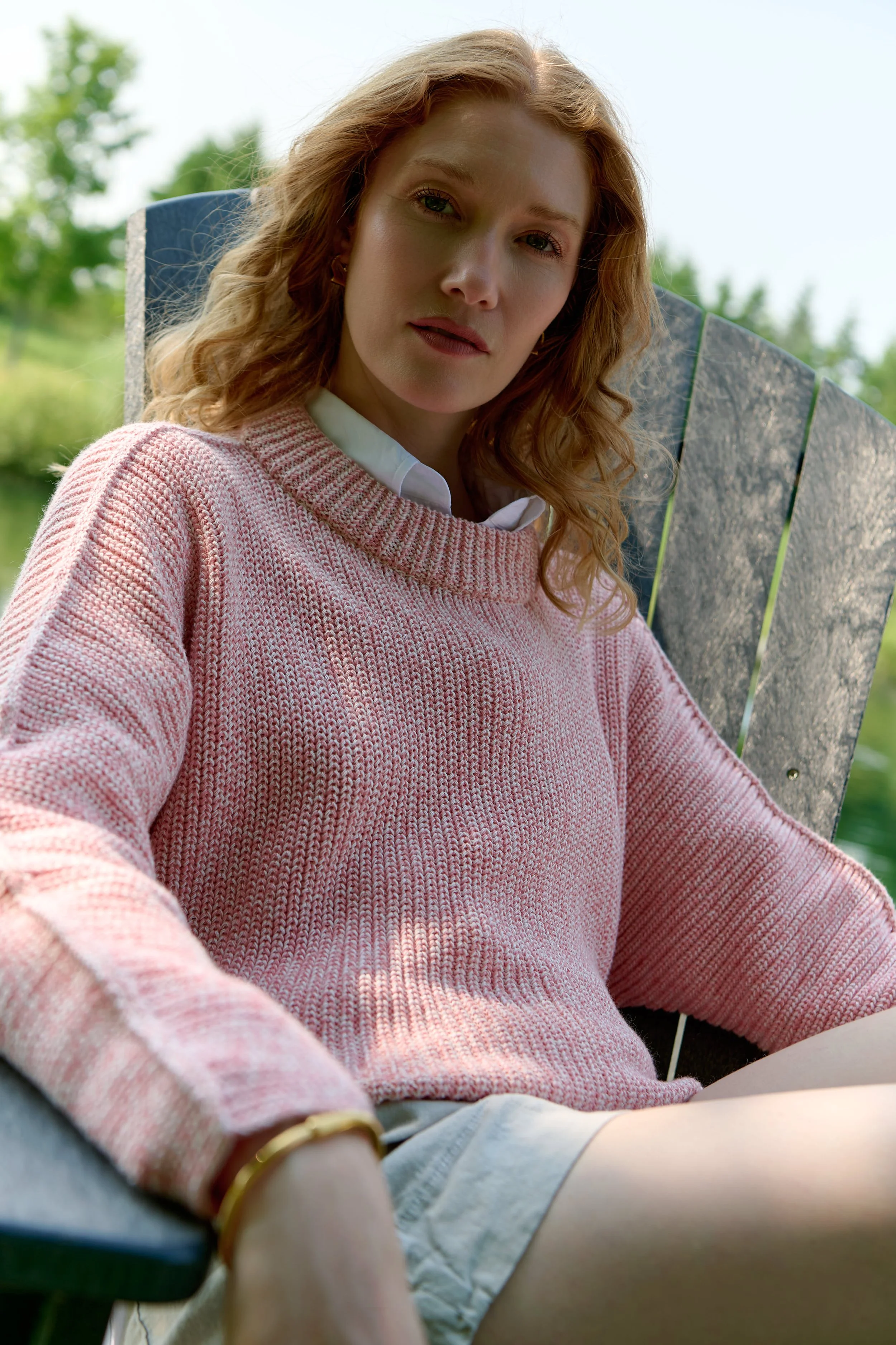 A woman with curly red hair sitting on a wooden chair outdoors, wearing a pink sweater over a white collared shirt and beige shorts.