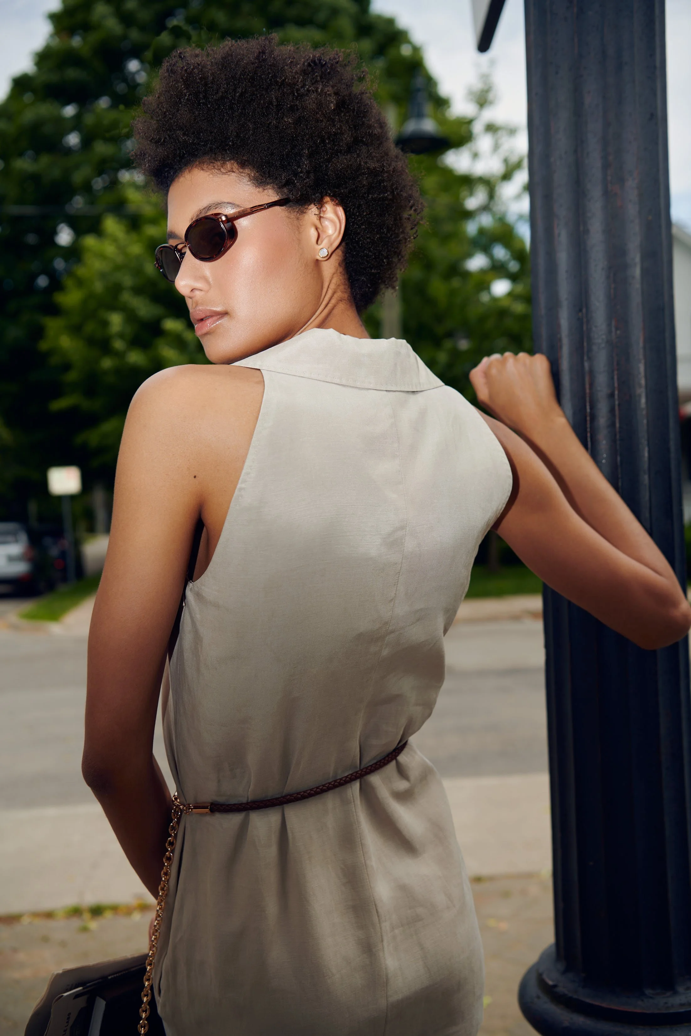 A woman with curly short hair wearing sunglasses and a sleeveless beige dress, standing outdoors during daytime near a black metal pole.