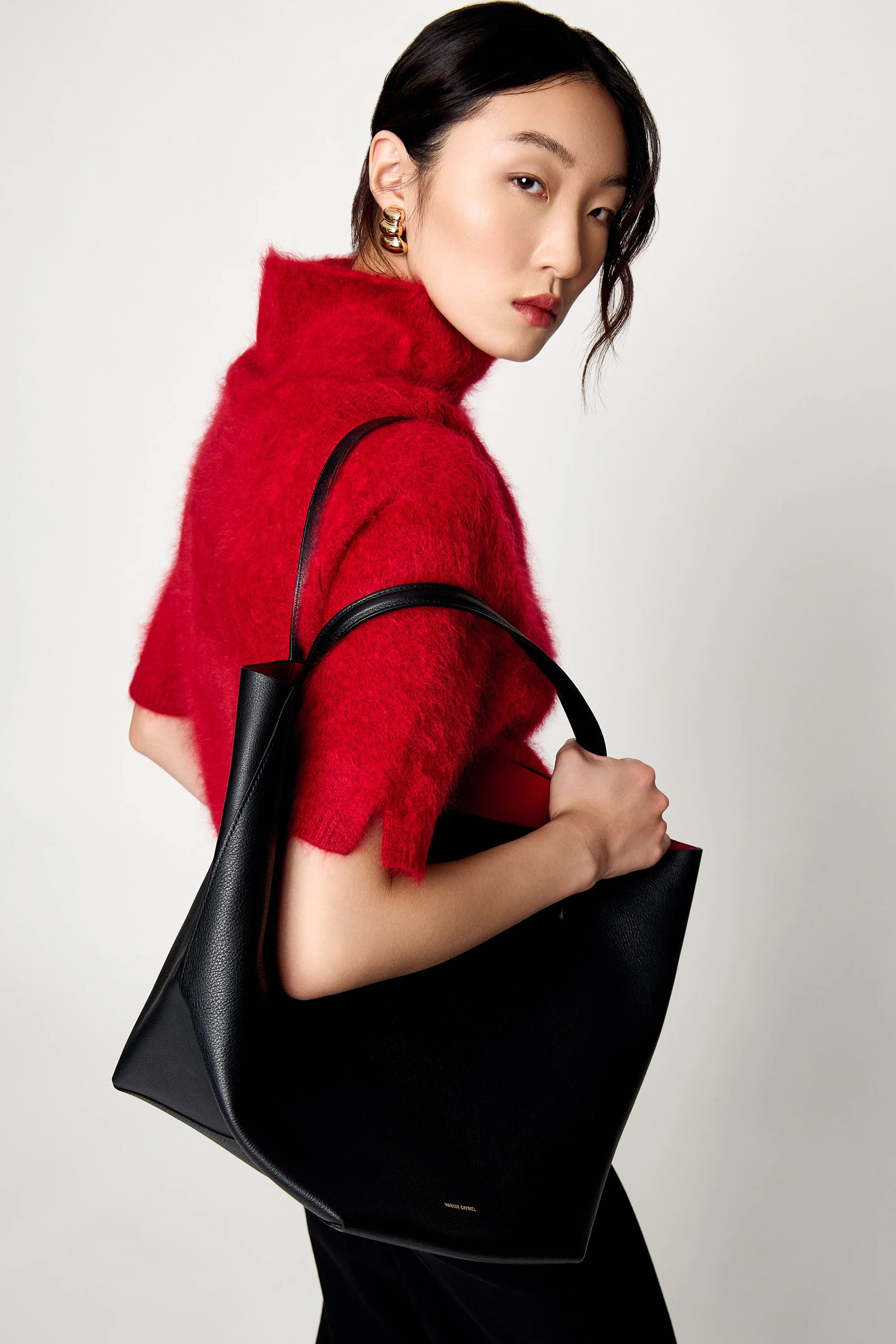 A woman with dark hair styled to the side, wearing a red fuzzy turtleneck sweater, gold hoop earrings, and holding a large black handbag over her shoulder, standing against a plain light gray background.