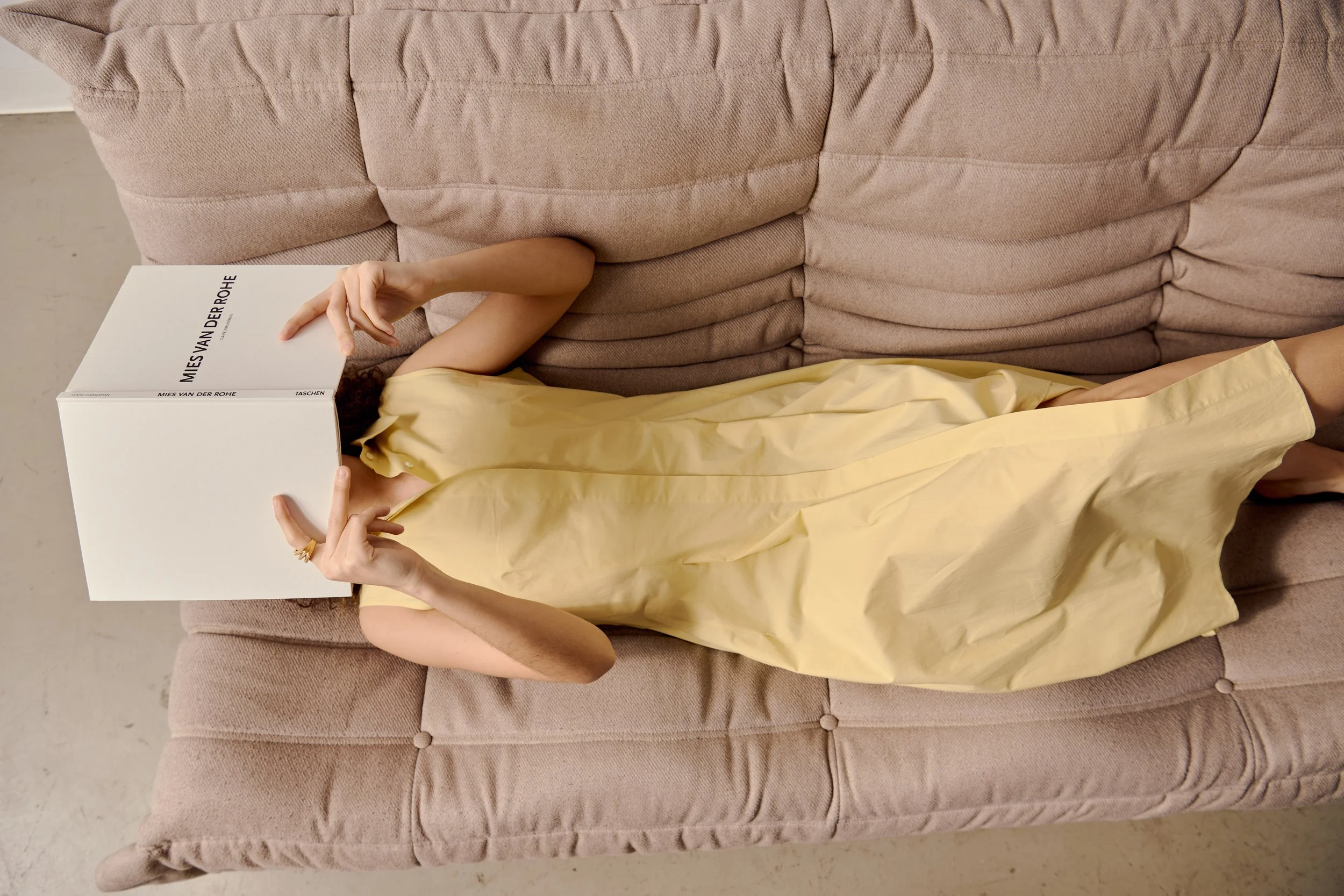 Person lying on a pink sofa, wearing a yellow dress, with a book covering their face, making a peace sign with one hand.