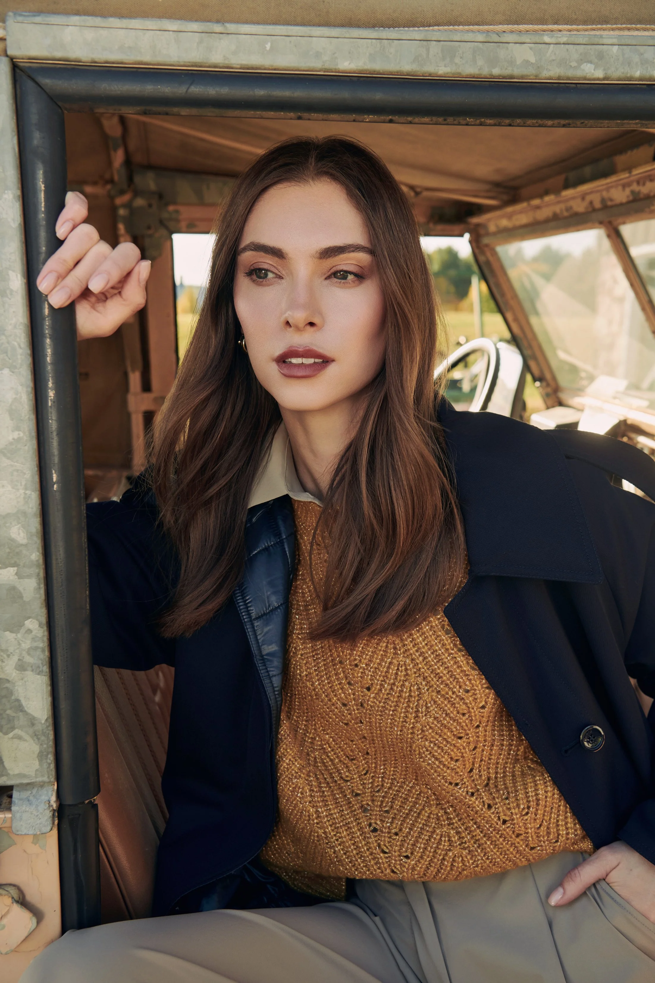 A woman with brown hair wearing a black jacket, beige blouse, and mustard sweater sitting in an old vehicle, looking thoughtfully into the distance.