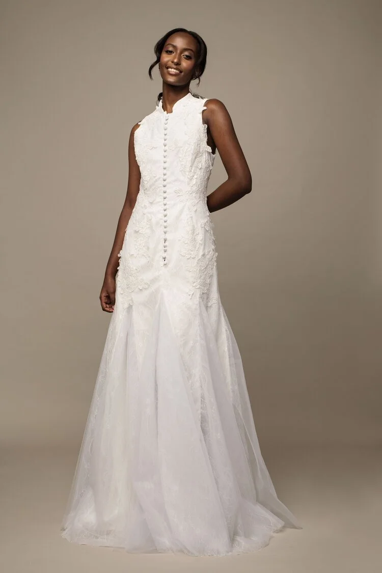 A woman in a sleeveless white lace wedding gown with buttons down the front, standing against a neutral background, smiling with one hand behind her back.