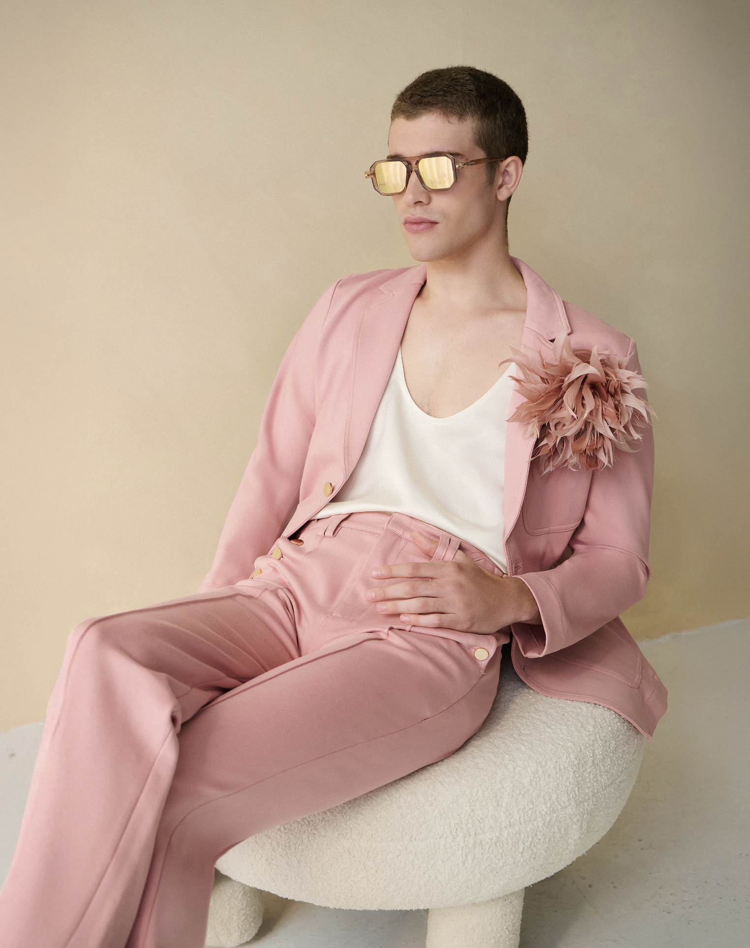 A young man with short brown hair wearing large yellow sunglasses, a pink suit with a floral embellishment on his shoulder, a white tank top, and pink pants, sitting on a textured white round chair against a beige background.