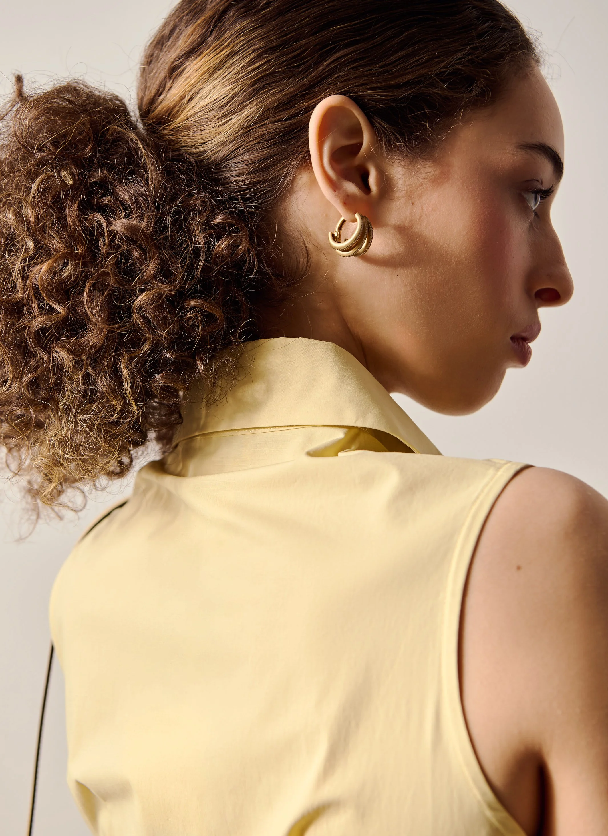 Close-up side profile of a woman with brown curly hair in a ponytail, wearing gold hoop earrings and a sleeveless yellow satin blouse against a plain background.
