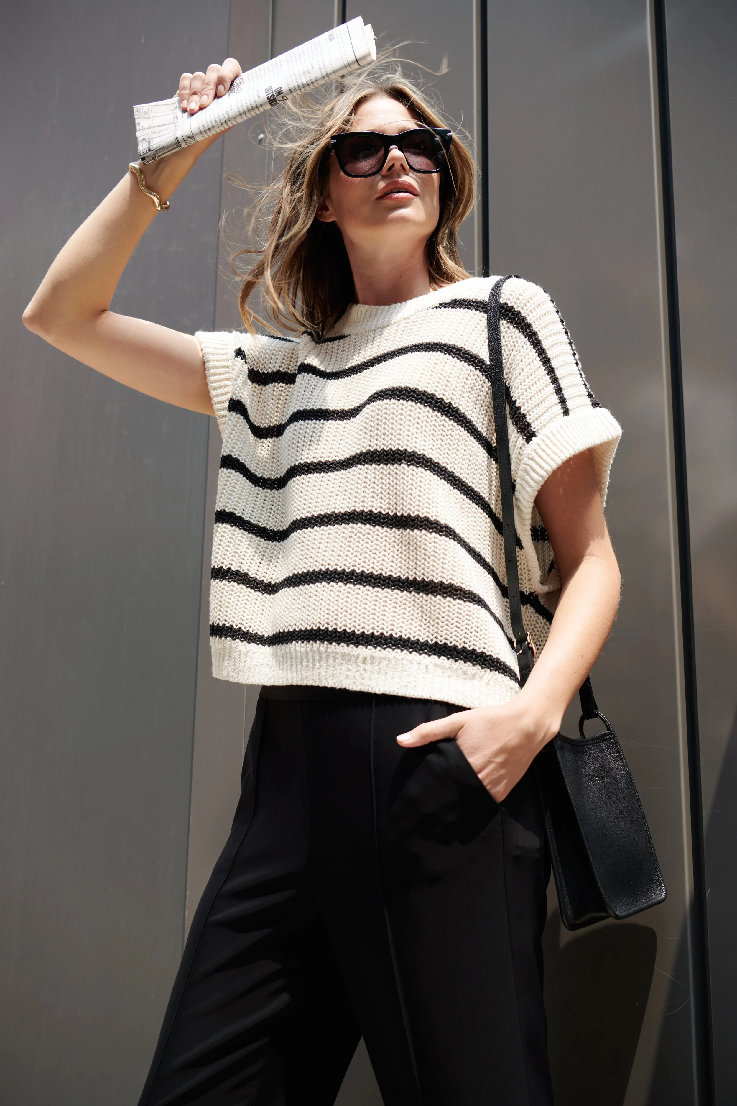 Stylish woman wearing sunglasses, a short-sleeve striped sweater, and black pants, holding a rolled-up newspaper above her head, standing outdoors against a metallic wall.