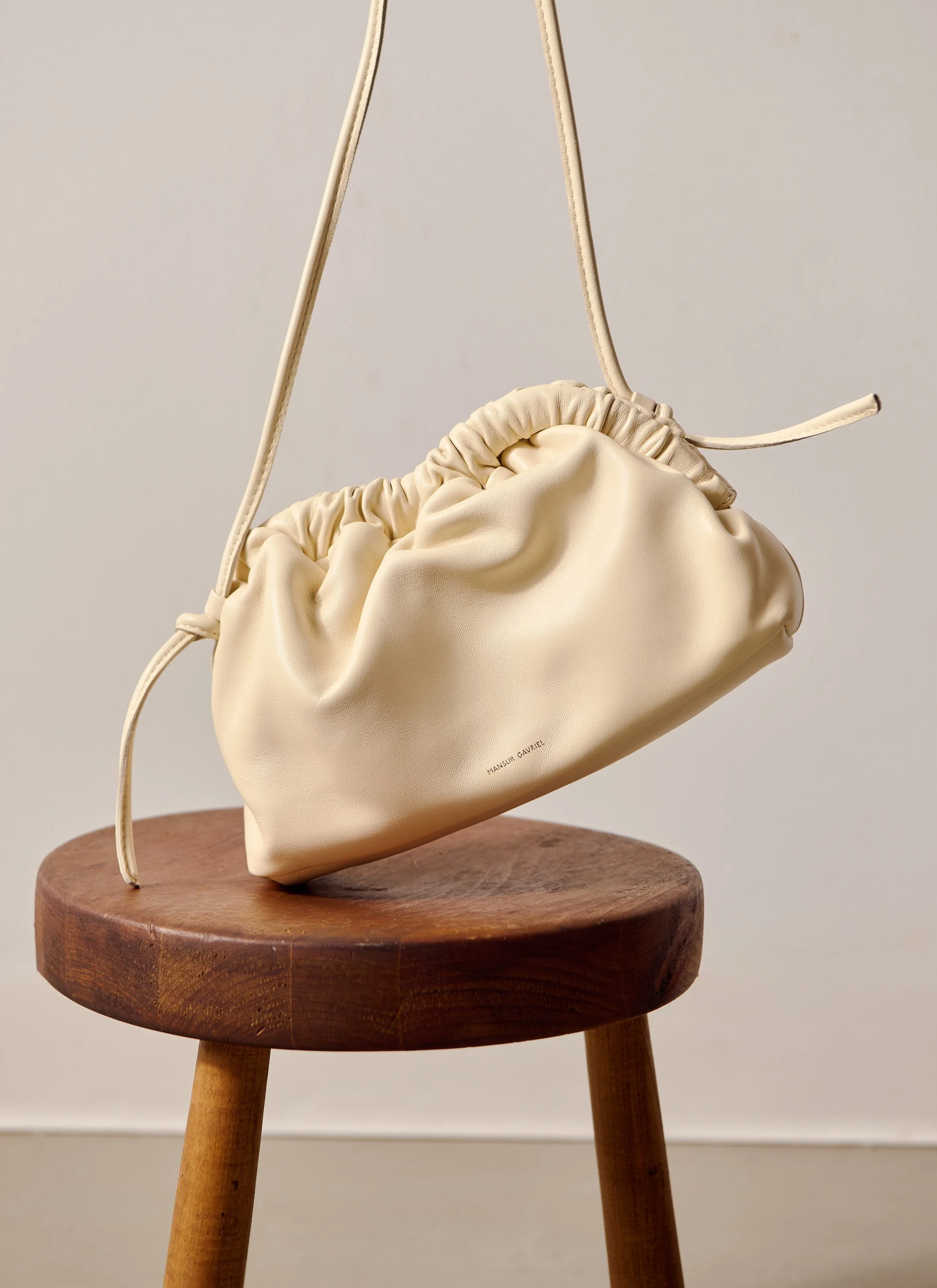 A cream-colored leather handbag with gathered detail hangs from a beige strap on a wooden stool against a neutral background.