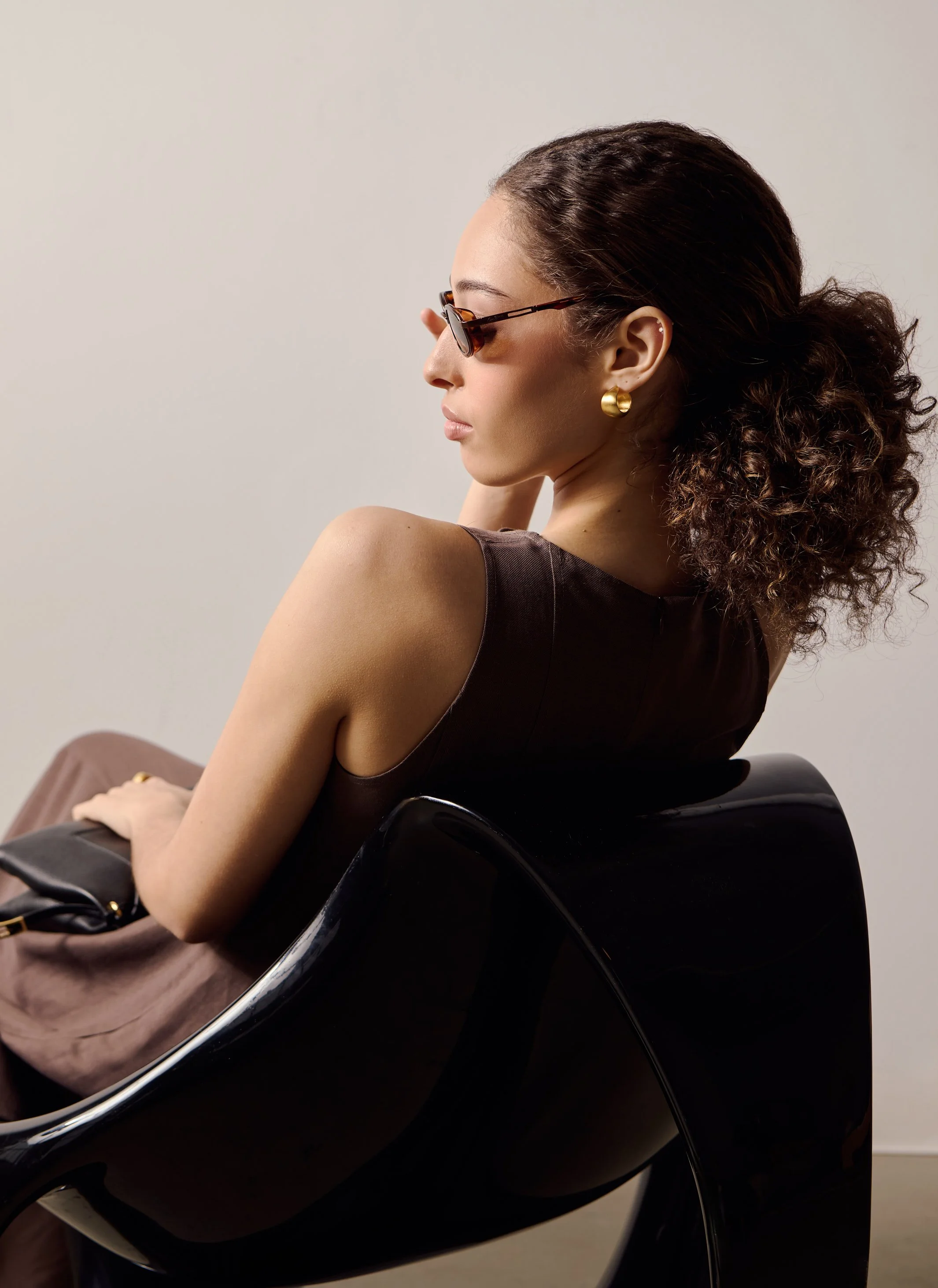 A woman with curly hair and gold earrings is sitting in a black chair, wearing sunglasses and a sleeveless dark top, against a plain light background.
