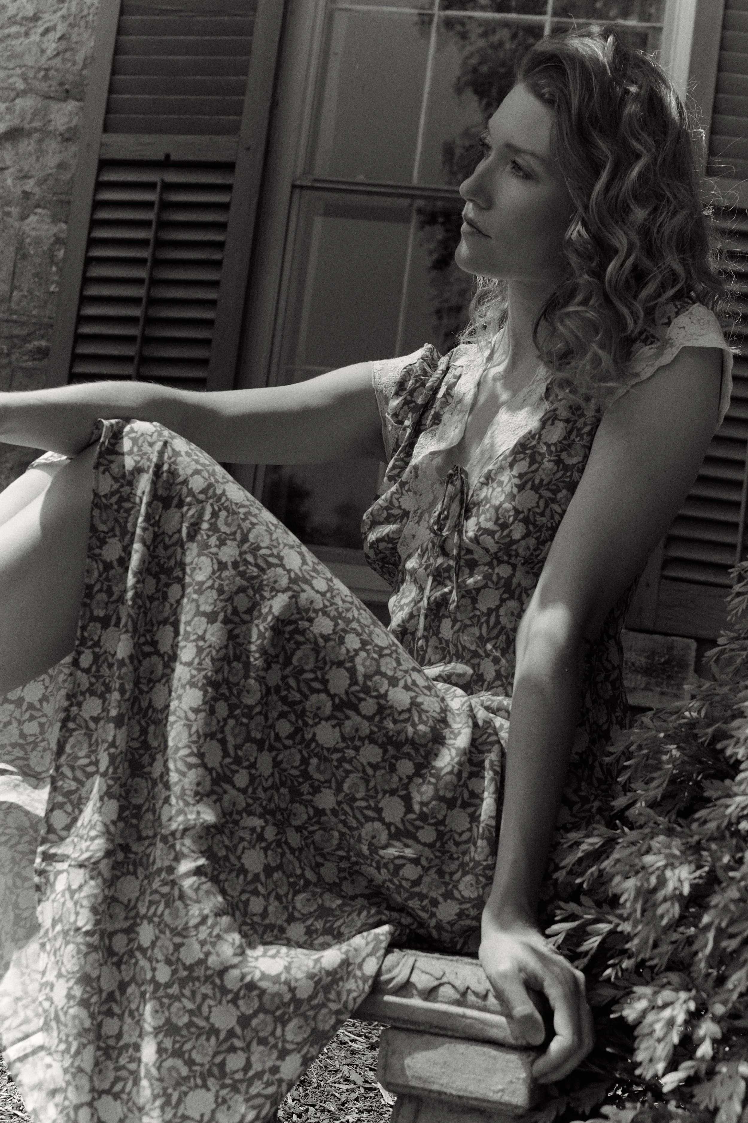 A woman with curly hair sitting on a bench outside near a window with shutters, wearing a floral dress and looking thoughtfully into the distance.