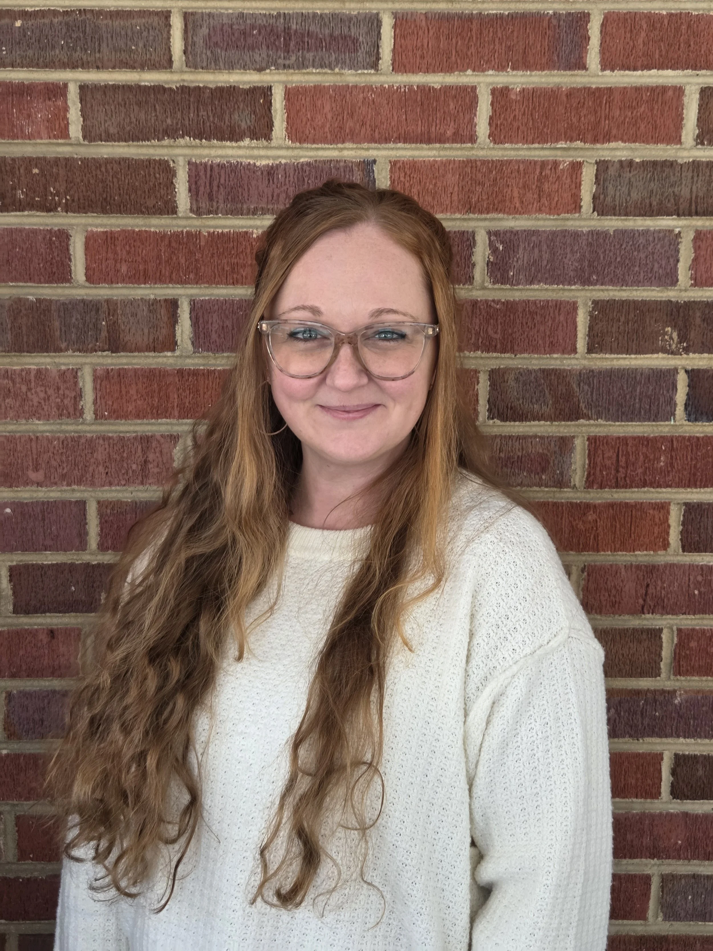 A woman with long wavy red hair and glasses standing against a red brick wall, wearing a white sweater.